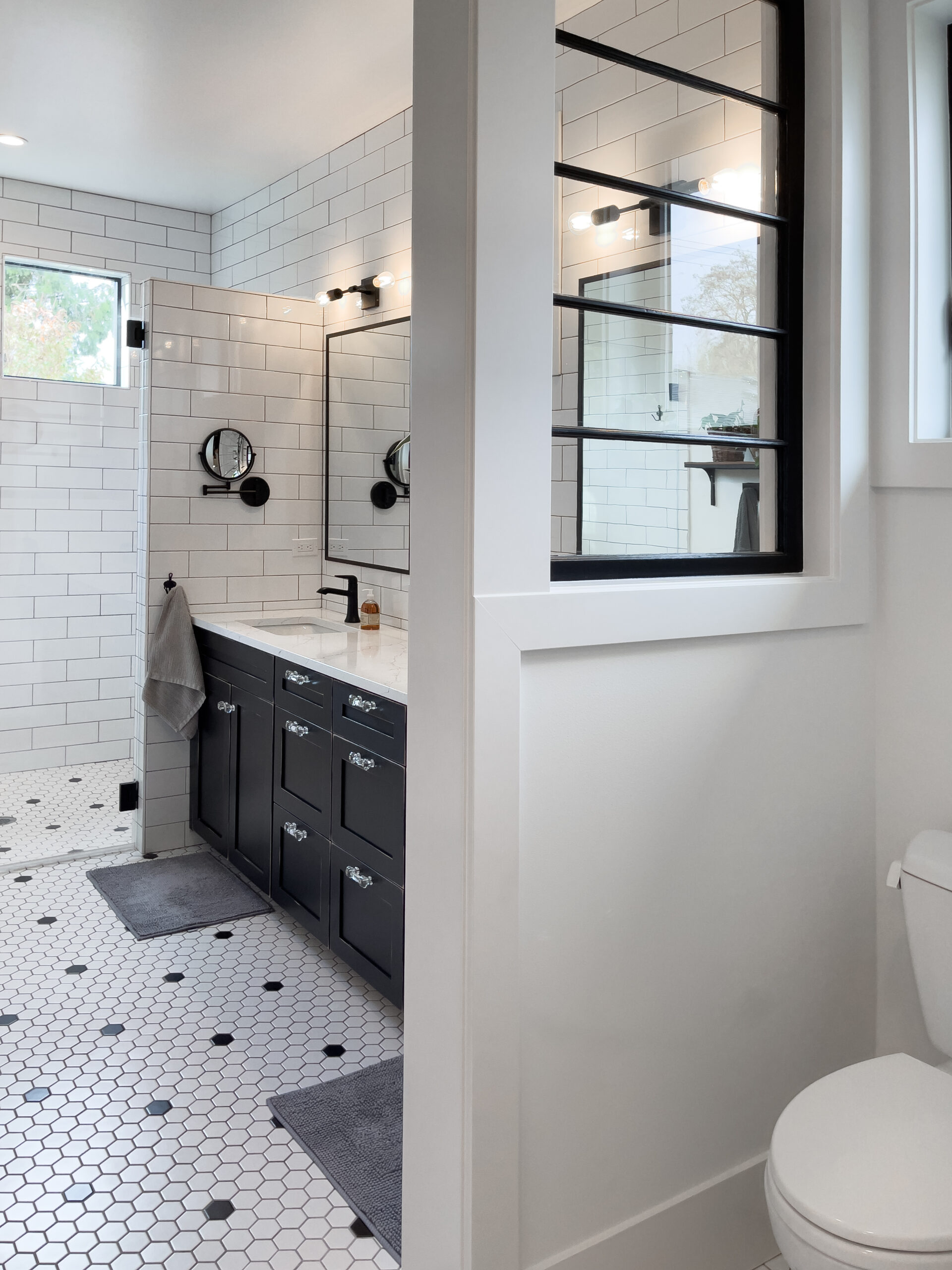 Modern bathroom with white subway tile walls, black vanity with double sinks, hexagonal floor tiles, two mirrors with lights above, a window, and a partial wall separating the toilet area.