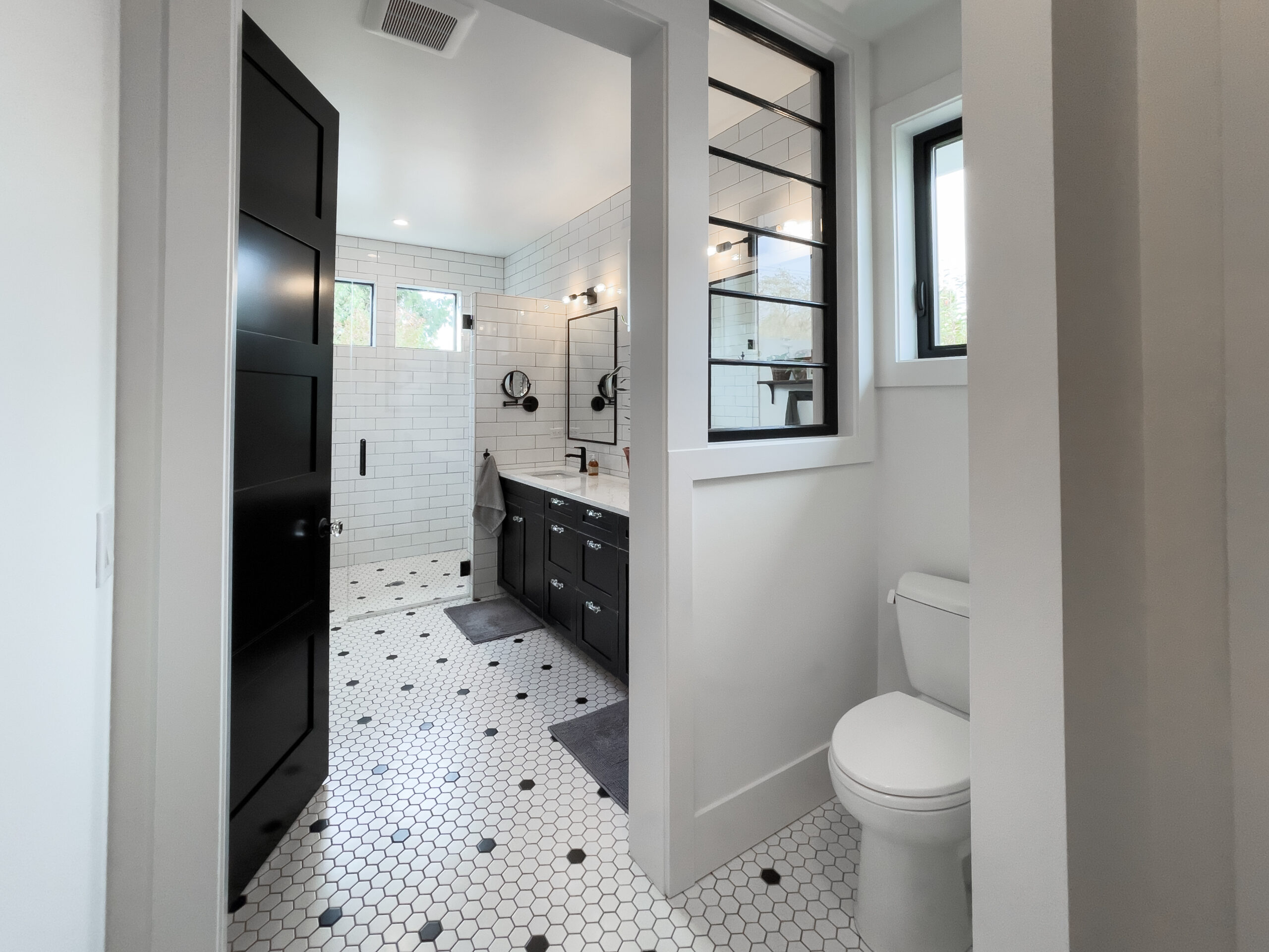 Modern bathroom with white hexagon tile floor, black vanity cabinets, double sinks with mirrors, white subway tile walls, and a window above the toilet. Black accents and natural light give the space a clean, contemporary look.