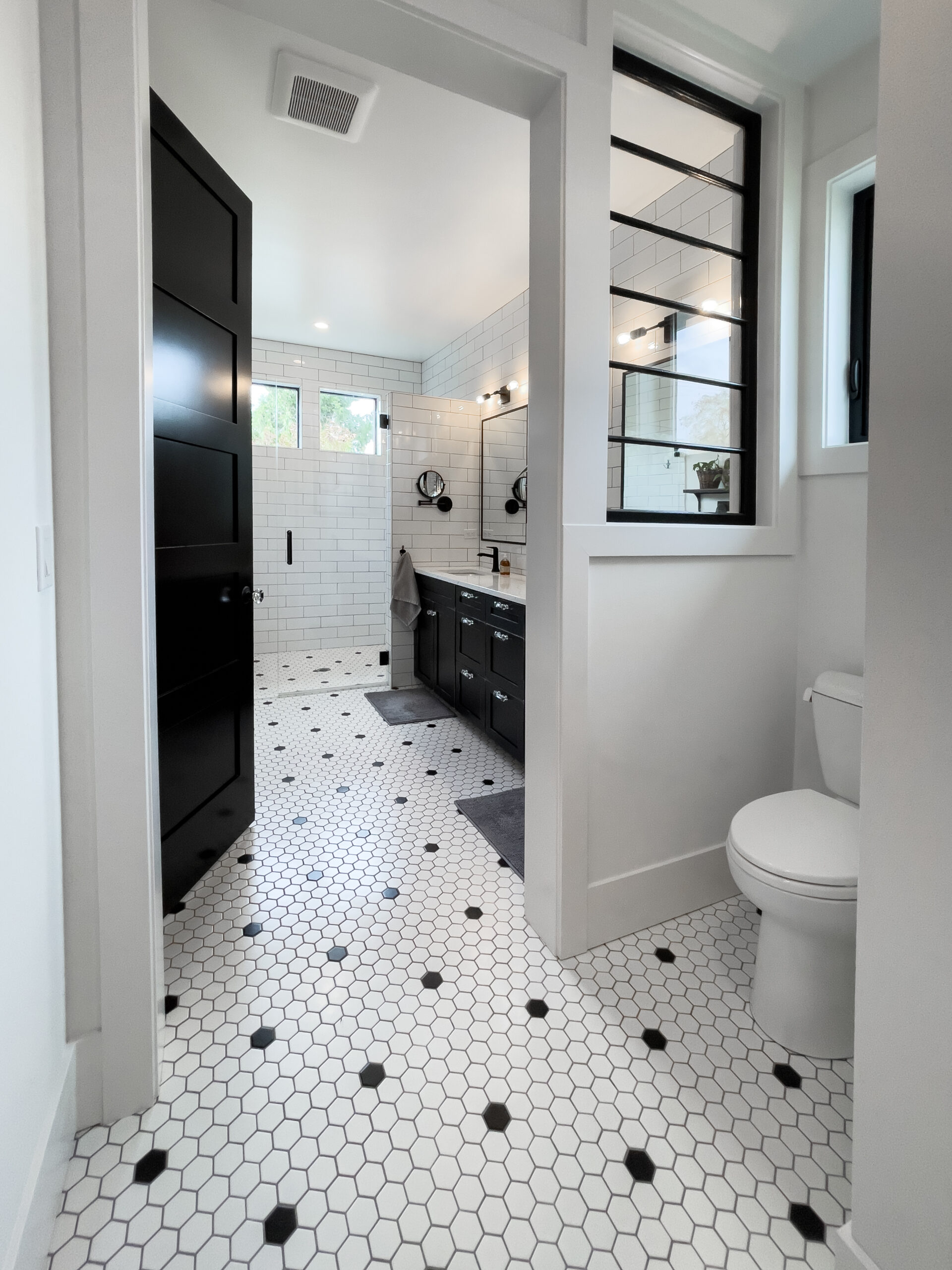 Modern bathroom with white hexagonal tile floor, black accents, double sink vanity with black cabinets, two mirrors, glass shower, and a toilet in a separate nook. White walls and black doors complete the clean design.