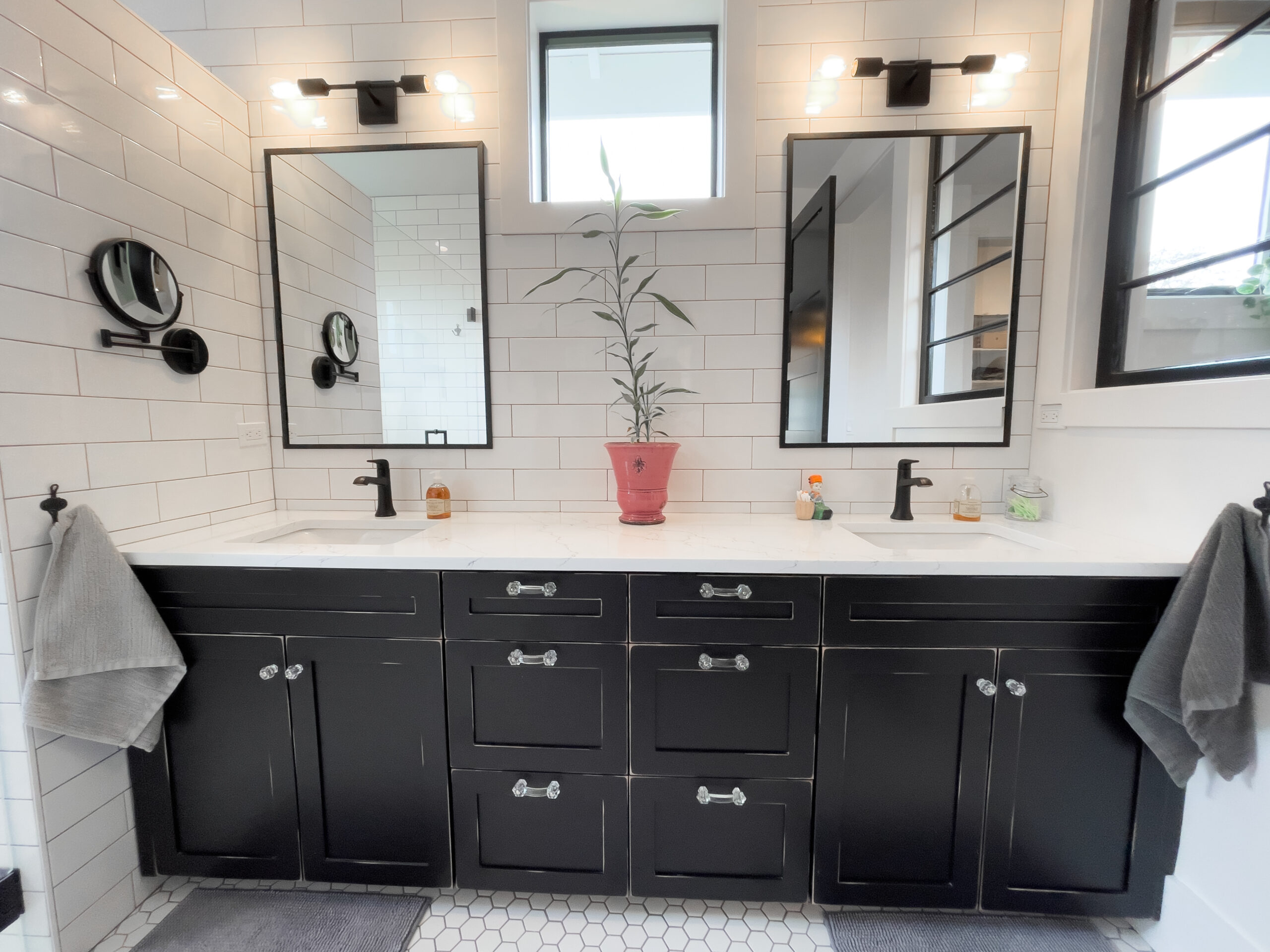 Modern bathroom with black cabinets, white countertops, two mirrors above dual sinks, black fixtures, gray towels, and a potted plant centered between the sinks. White subway tiles and hexagonal floor tiles complete the look.