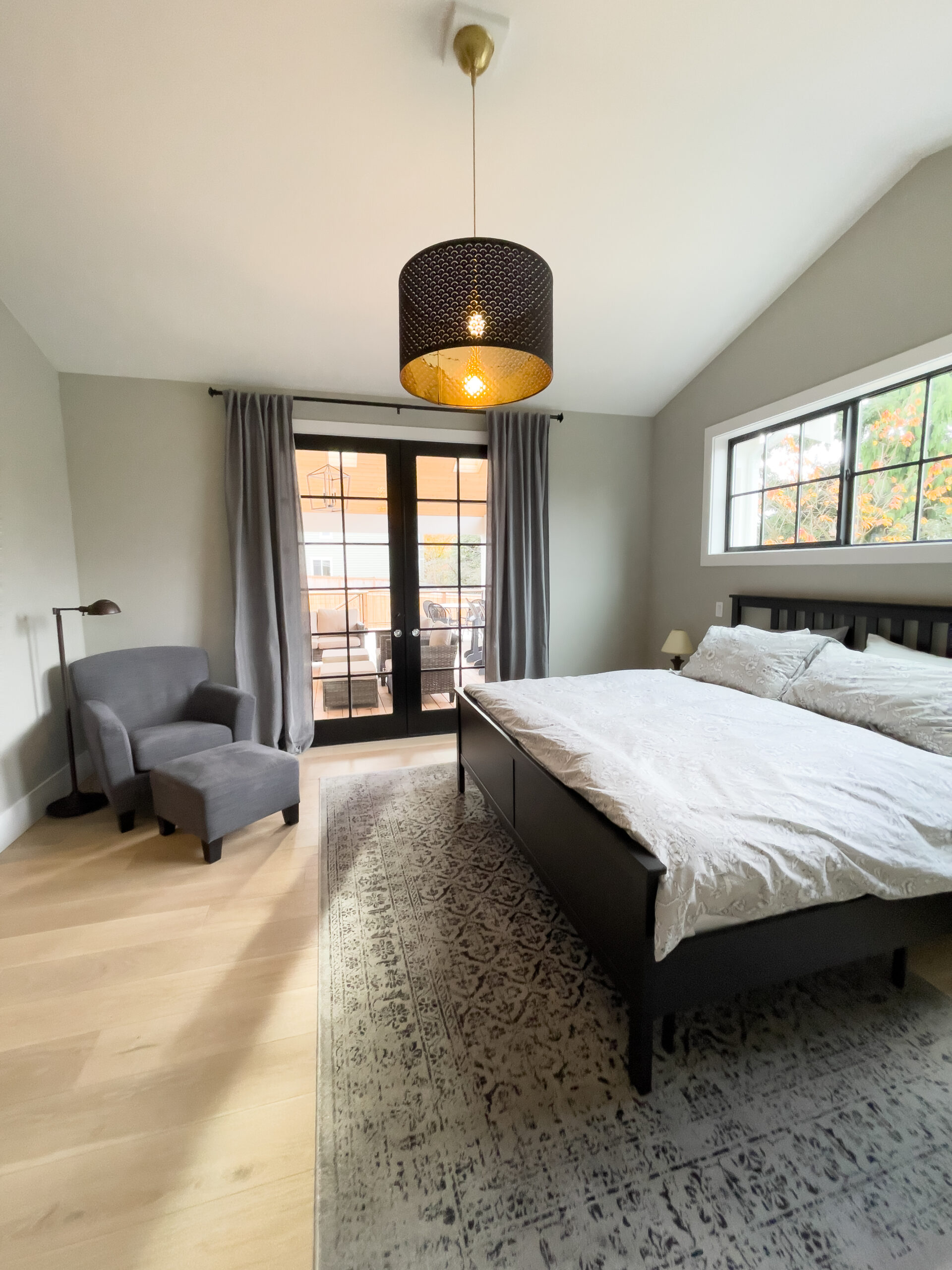 Modern bedroom with a large bed, armchair, floor lamp, and rug. French doors with dark curtains open to a balcony. A large window lets in natural light. Black pendant light hangs from the ceiling.