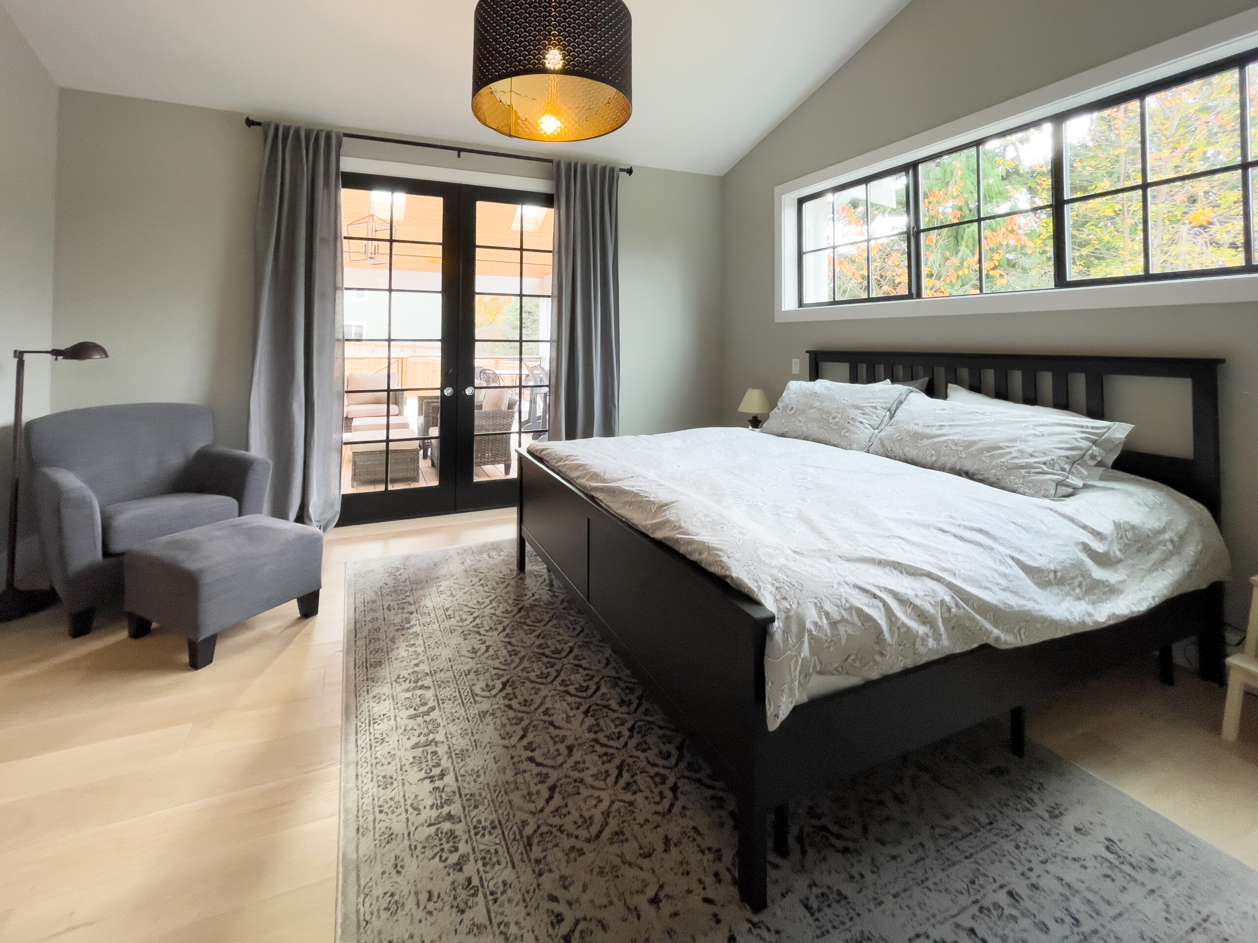 A modern bedroom with a large bed, white bedding, a patterned rug, an armchair, and floor lamp. French doors with gray curtains and a wide window let in natural light. The walls are light gray and the floor is light wood.
