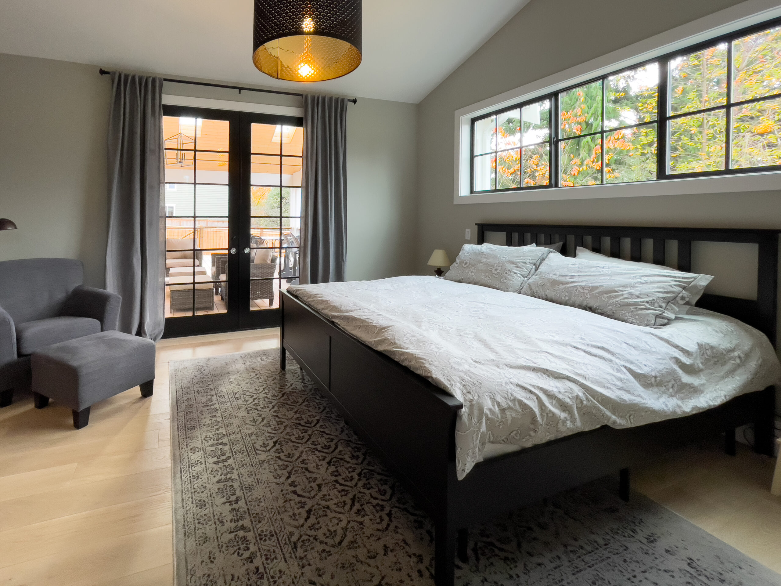 A modern bedroom with a large bed, patterned rug, gray walls, armchair, and double glass doors leading to a patio. A window lets in natural light and trees are visible outside.