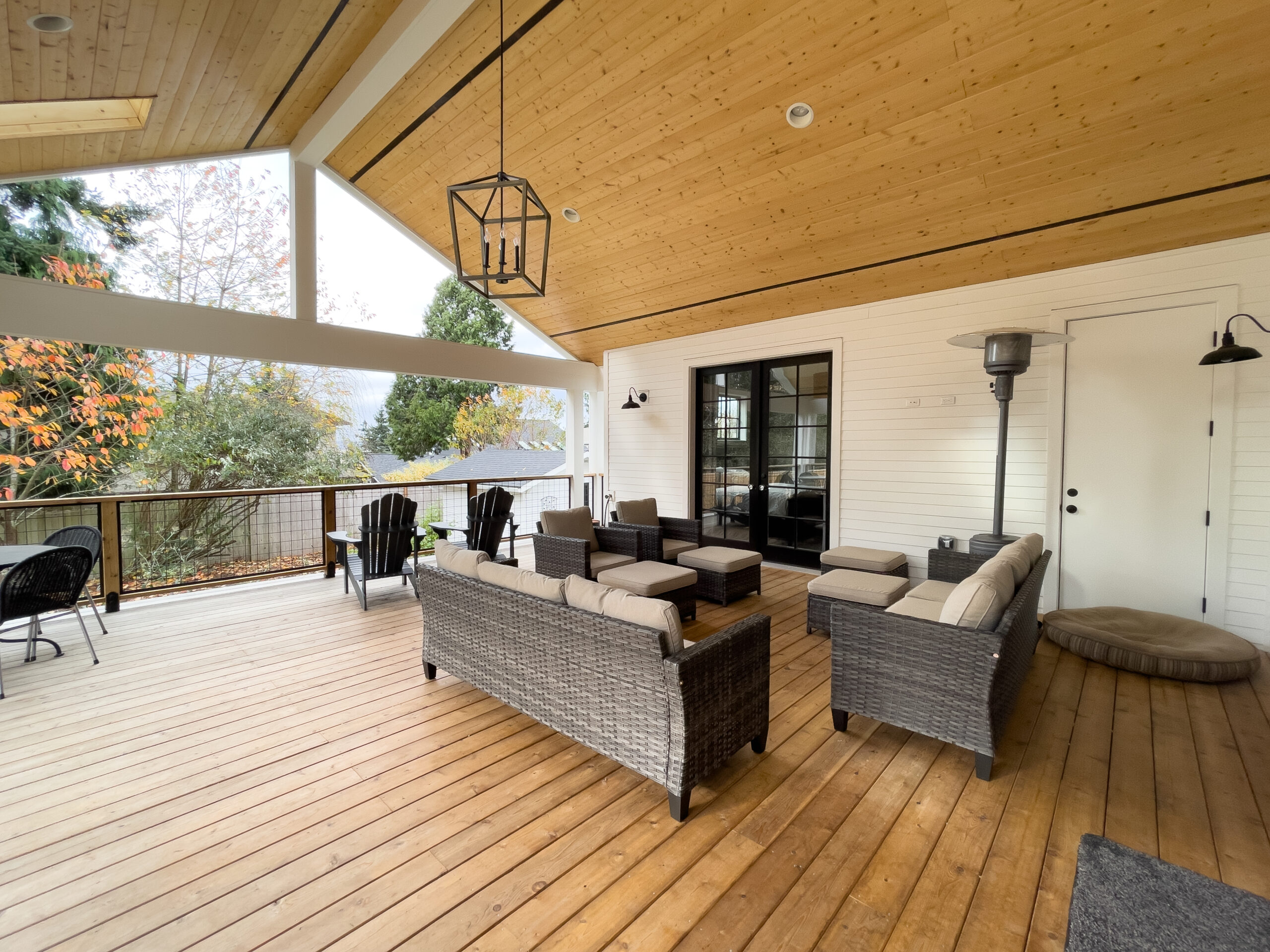 A covered outdoor patio with wooden flooring and ceiling, featuring wicker sofas with beige cushions, a black-framed glass door, and a view of trees and houses in the background.