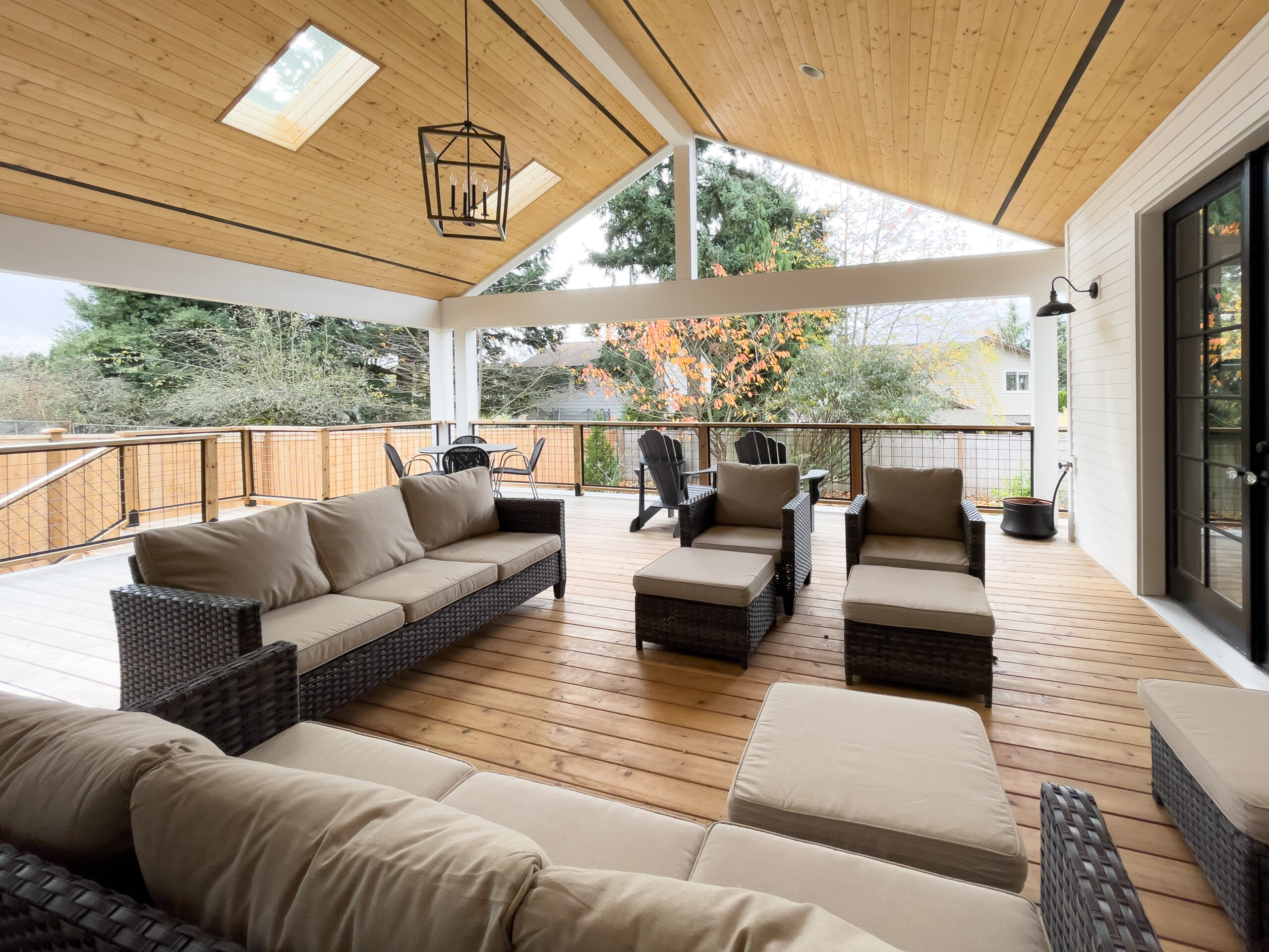 A spacious covered patio with wooden flooring, wicker furniture with beige cushions, skylights in the wooden ceiling, and a view of trees and houses outside.