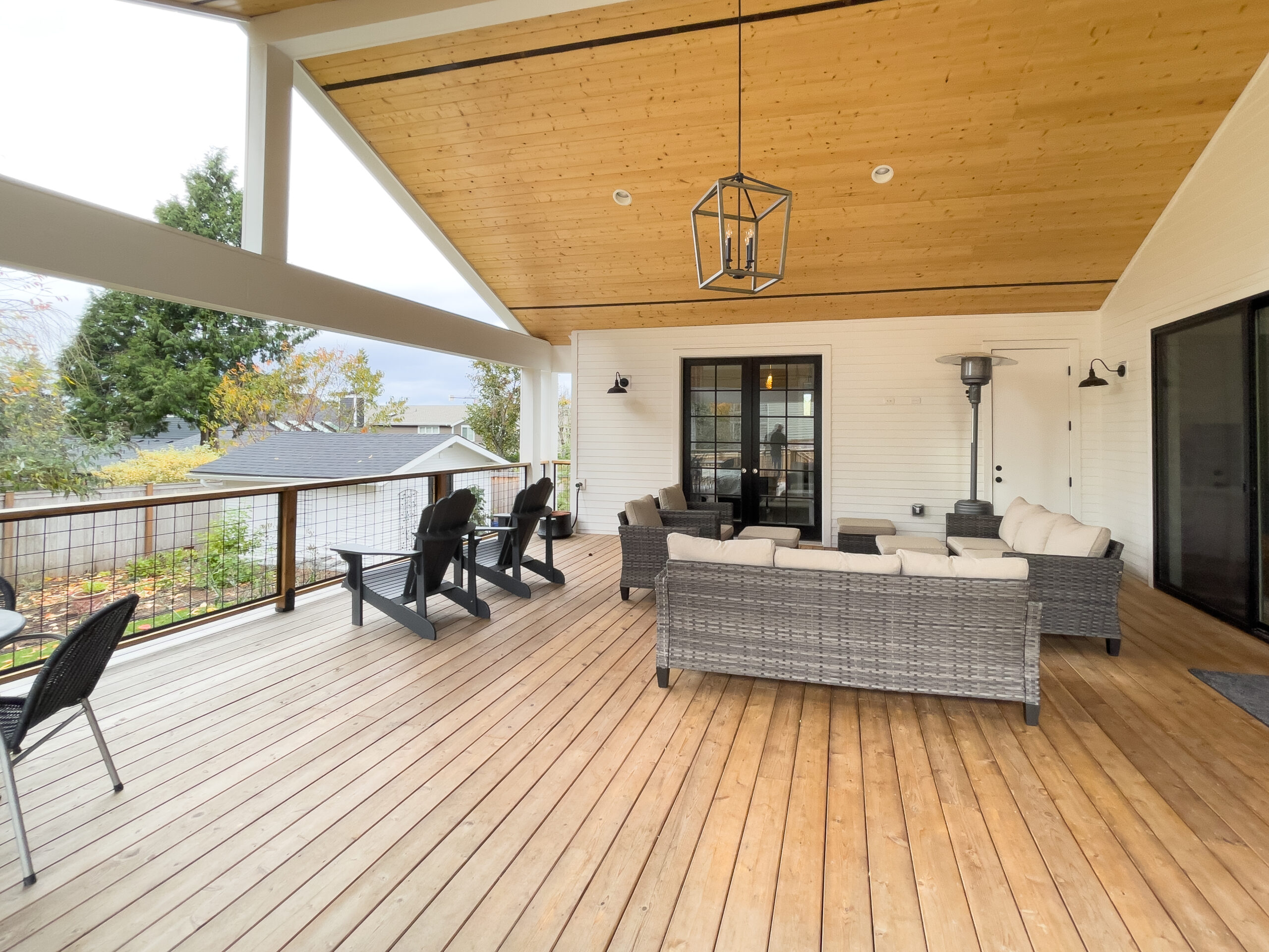 A spacious covered patio with a wooden deck, wicker sofas, armchairs, a heater, and a lantern-style ceiling light. The area overlooks a garden and neighboring house, with trees visible in the background.