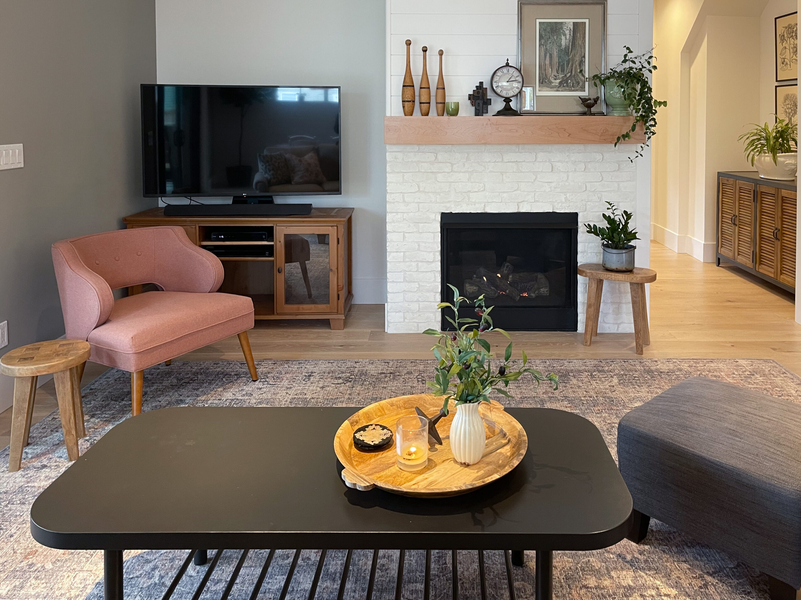 A cozy living room with a fireplace, TV, pink chair, wooden furniture, and a black coffee table topped with a tray, plant, candle, and pottery. A patterned rug covers the floor and decor accents the mantle and walls.