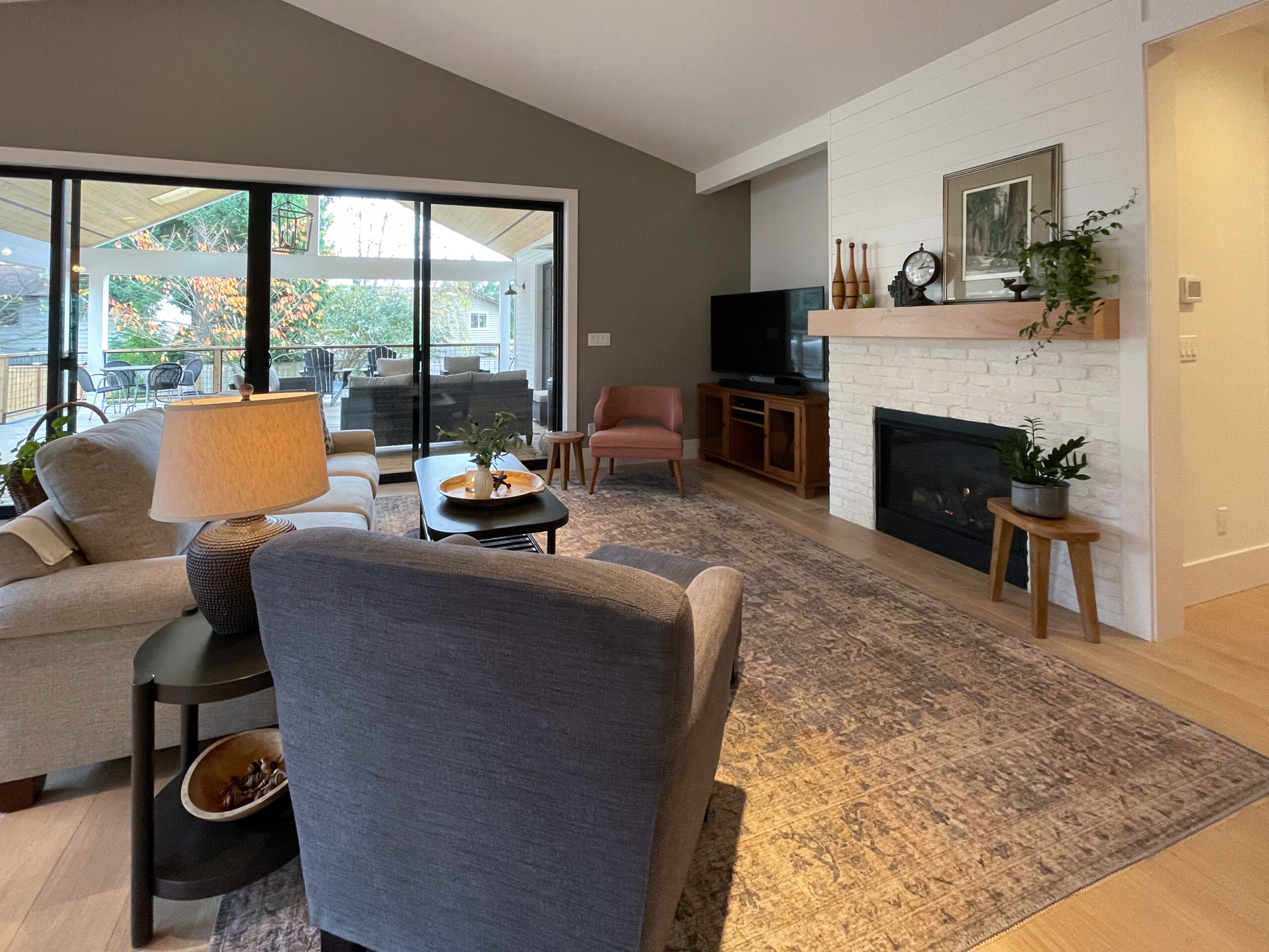 A cozy living room with neutral furniture, a large area rug, a fireplace with a mantel, plants, a TV, and sliding glass doors opening to a covered patio with outdoor seating and greenery visible outside.