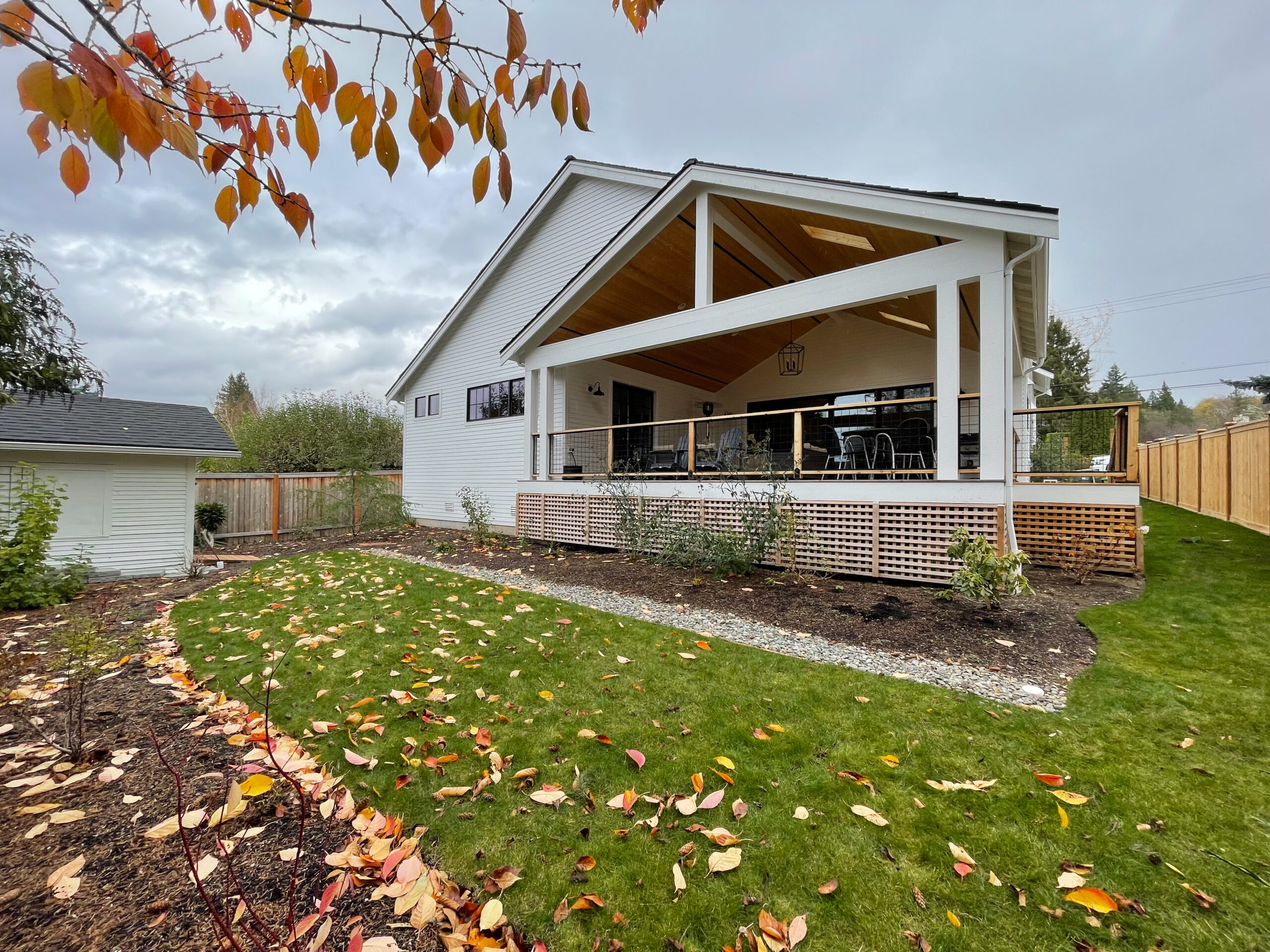 A modern white house with a large covered patio and wooden railings, surrounded by a green lawn and autumn leaves. Trees with orange foliage are in the foreground, and a wooden fence lines the yard.