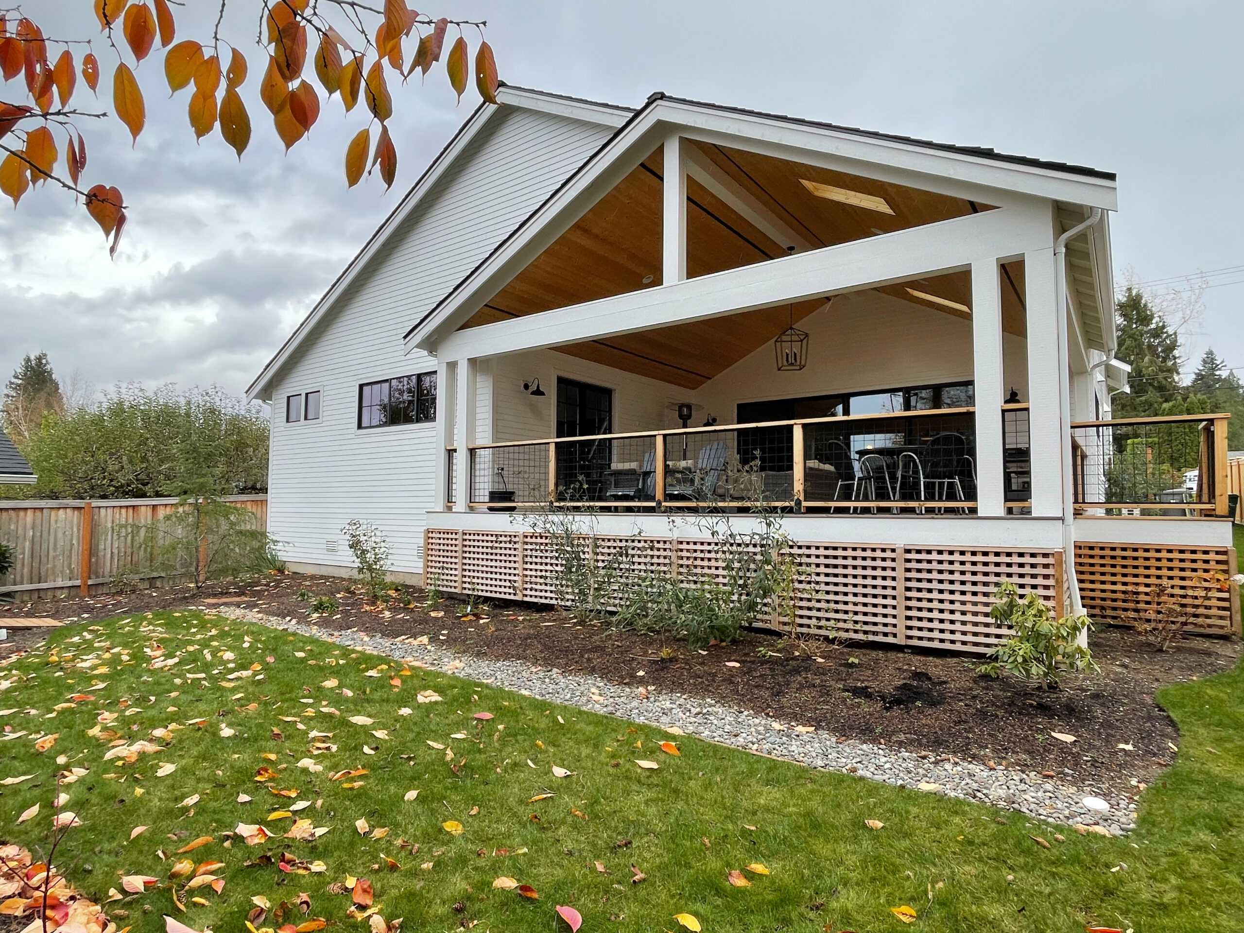 A modern white house with a large covered back porch featuring wooden accents and black railing, overlooking a green lawn with scattered autumn leaves and a garden bed along the edge.