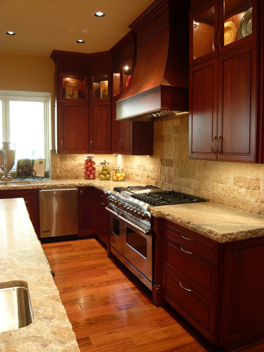 A warm, elegant kitchen with dark wood cabinets, stainless steel appliances, a large oven and stove, beige stone countertops, under-cabinet lighting, and a window letting in natural light.