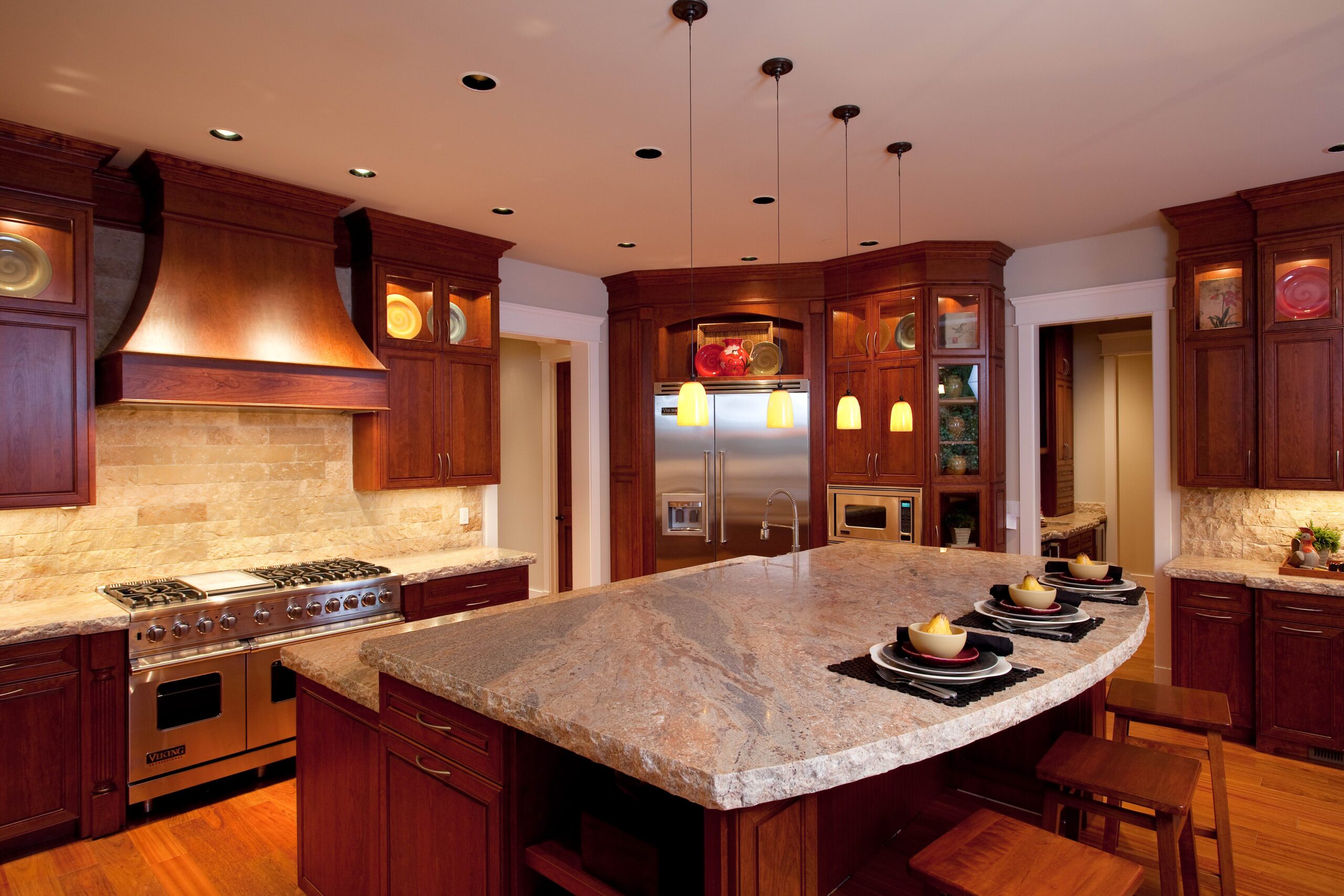 Spacious kitchen with dark wood cabinets, stainless steel appliances, a large marble island with three place settings, pendant lights, and beige stone backsplash.