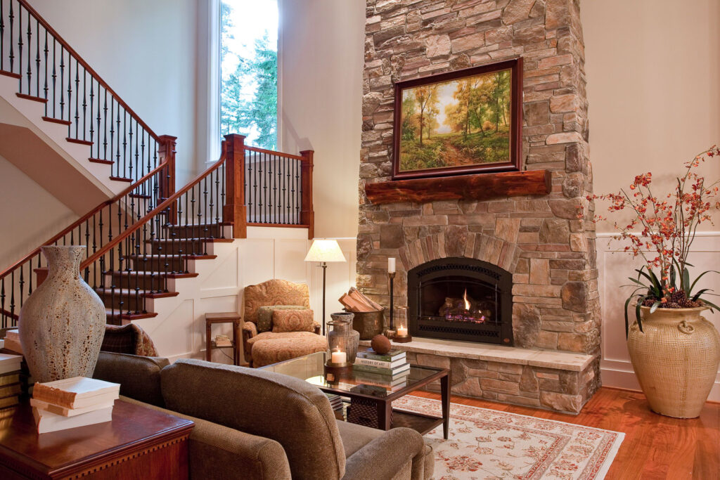 A cozy living room with a stone fireplace, artwork above the mantel, plush seating, a patterned rug, large vases, books, soft lighting, and a staircase with wooden railings to the left. Tall windows bring in natural light.