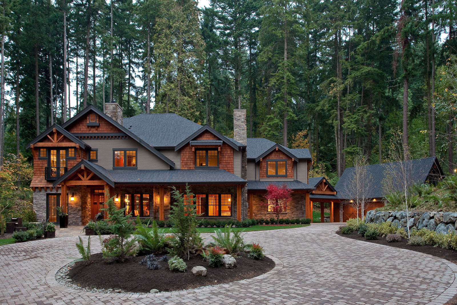 Large Lodge Style-11 A large, rustic-style house with wooden and stone accents, lit warmly from within, sits surrounded by tall trees and lush landscaping. A circular brick driveway leads to the entrance.