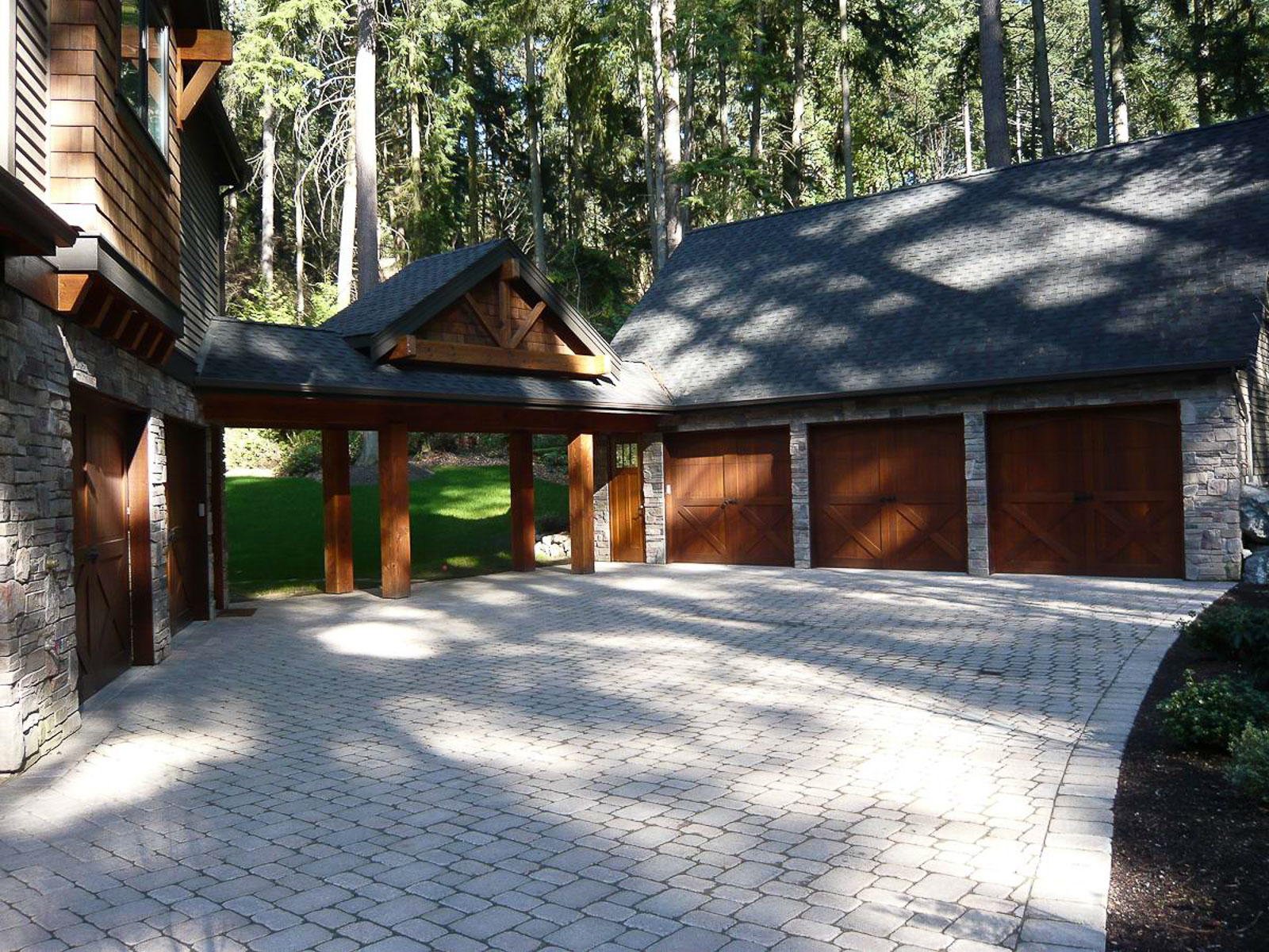 Large Lodge Style-13 Large wooden house with stone accents, featuring a three-car garage with brown wooden doors and a spacious, light-colored cobblestone driveway surrounded by trees and greenery.