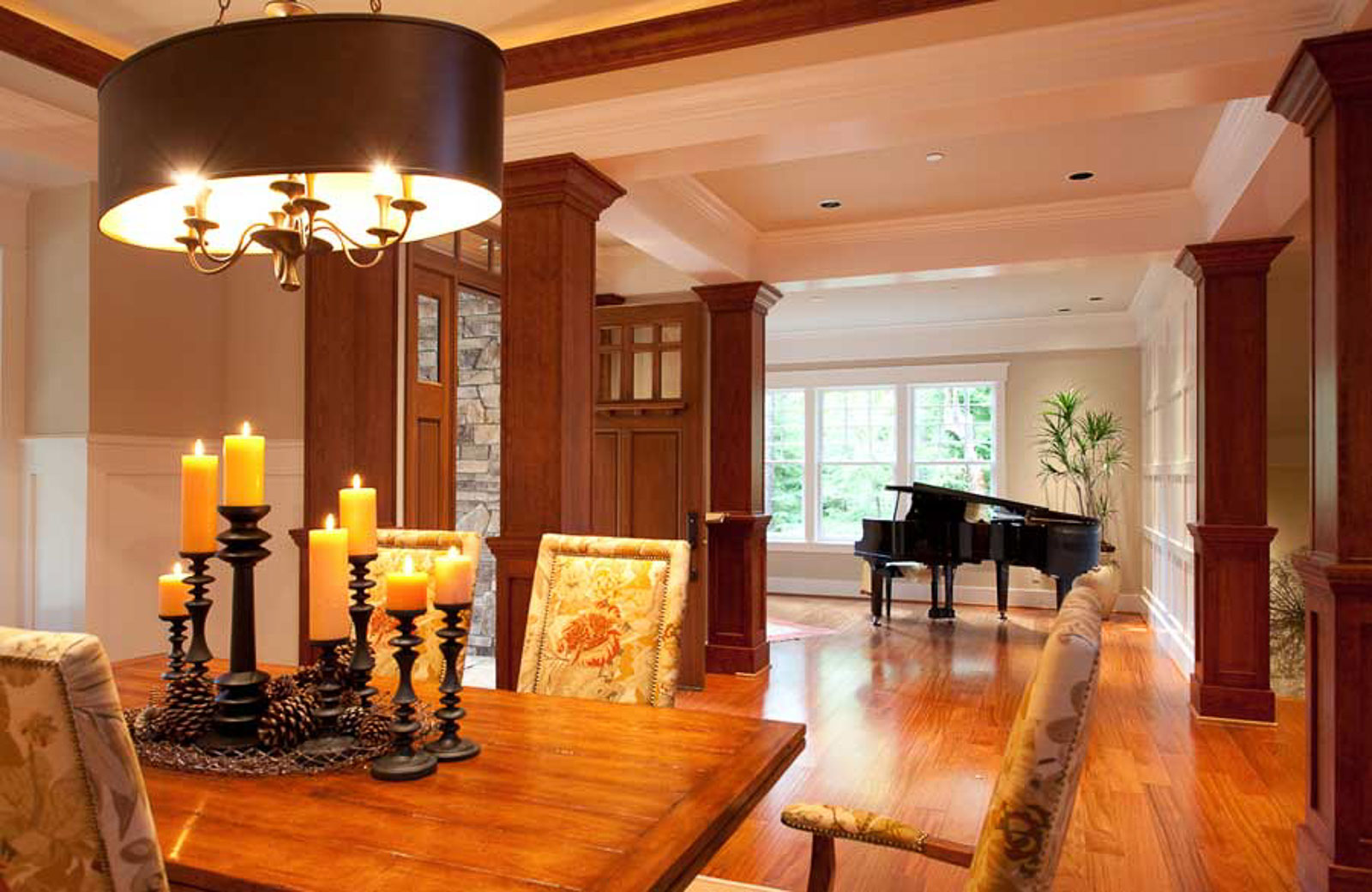 Large Lodge Style-3 A warmly lit dining room with a wooden table, floral upholstered chairs, and a black chandelier with multiple candles. In the background, a black grand piano sits near large sunlit windows.