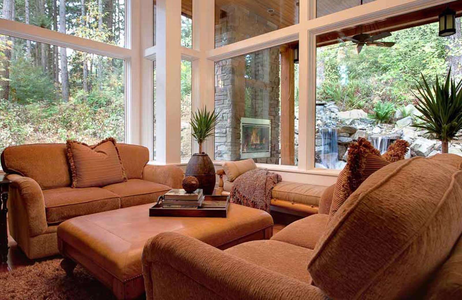 Large Lodge Style-4 A cozy living room with brown sofas and ottoman, large floor-to-ceiling windows showing a forest and waterfall outside, and indoor plants on small tables. A fireplace is visible on a stone wall outside.
