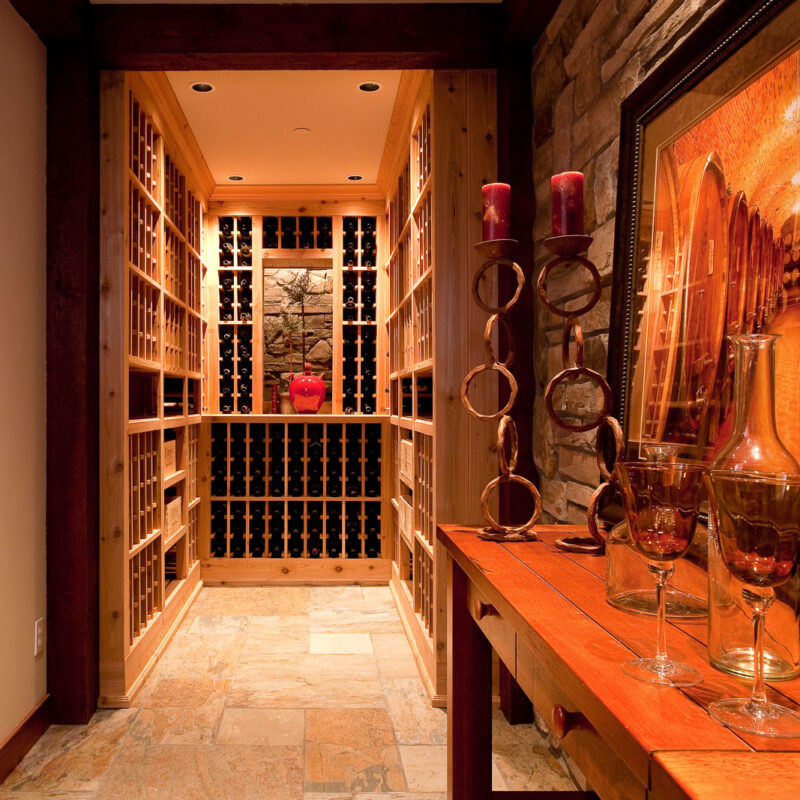 A cozy wine cellar with wooden racks filled with bottles, stone tile flooring, and a side table holding wine glasses, candles, and a framed wine-themed painting. Warm lighting creates an inviting atmosphere.