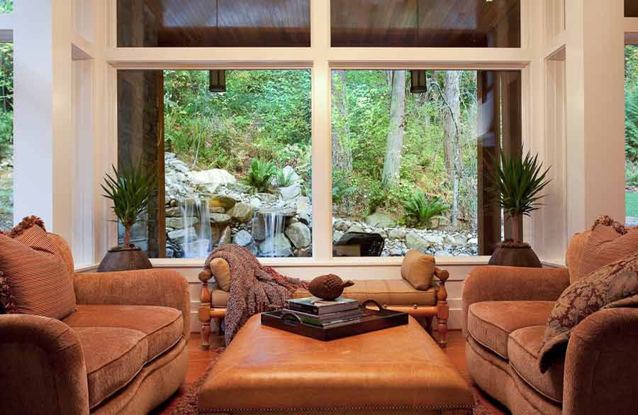 A cozy living room with two brown armchairs and a large ottoman faces a window with a view of a lush green garden, rocks, and a small waterfall. Two potted plants sit beside the window.