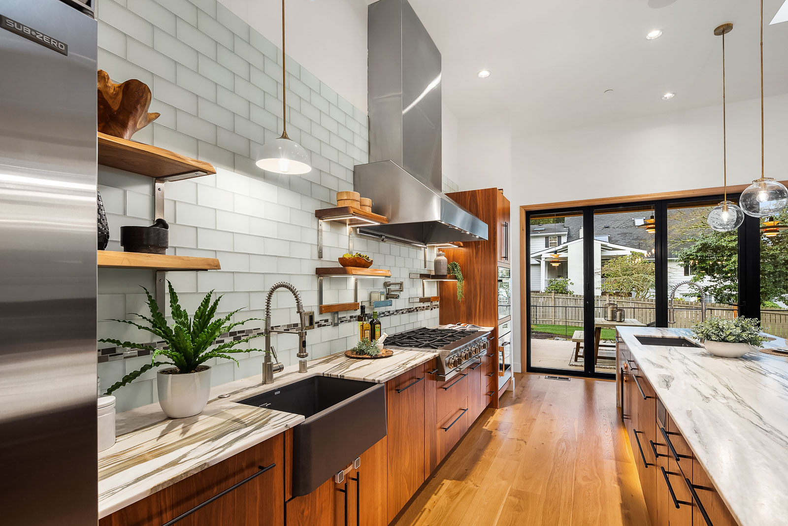 Luxury Mercer Island Mid-Century Modern-11 Modern kitchen with wood cabinets, marble countertops, a large farmhouse sink, stainless steel appliances, open shelving, and white subway tile backsplash; glass doors open to a backyard with a lawn and trees.