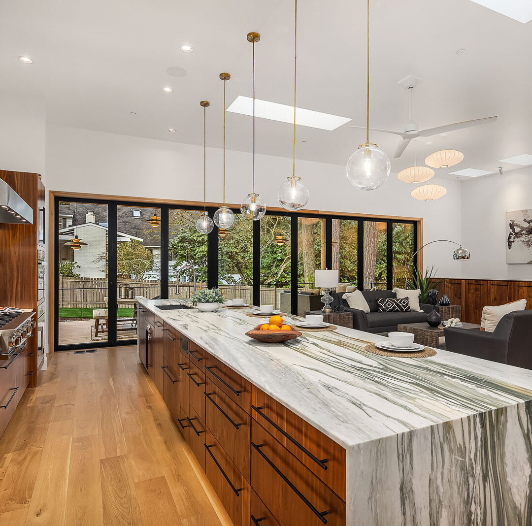 Modern kitchen with a long marble island, wooden cabinets, pendant lights, and open shelving. The space opens to a bright living area with large sliding glass doors, wood floors, and outdoor views.