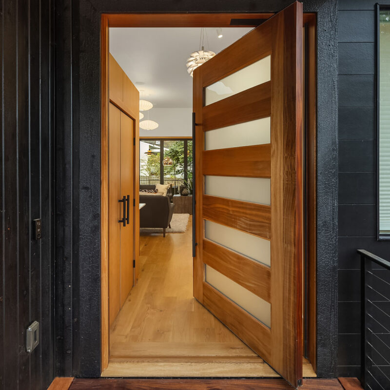 A modern wooden front door with frosted glass panels is partially open, revealing a bright interior with wood floors, contemporary lighting, and a view through to a living room and large windows.