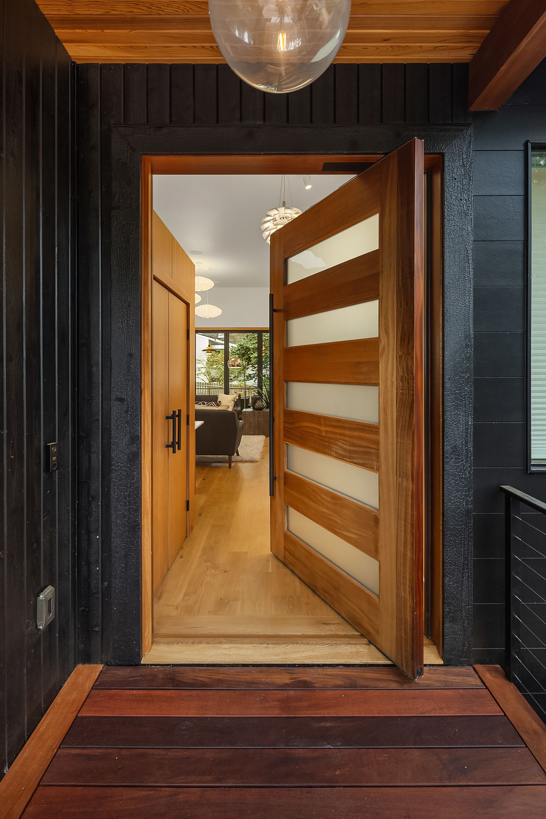 Luxury Mercer Island Mid-Century Modern-17 A modern wooden front door with frosted glass panels is partially open, revealing a bright interior with wood floors, contemporary lighting, and a view through to a living room and large windows.