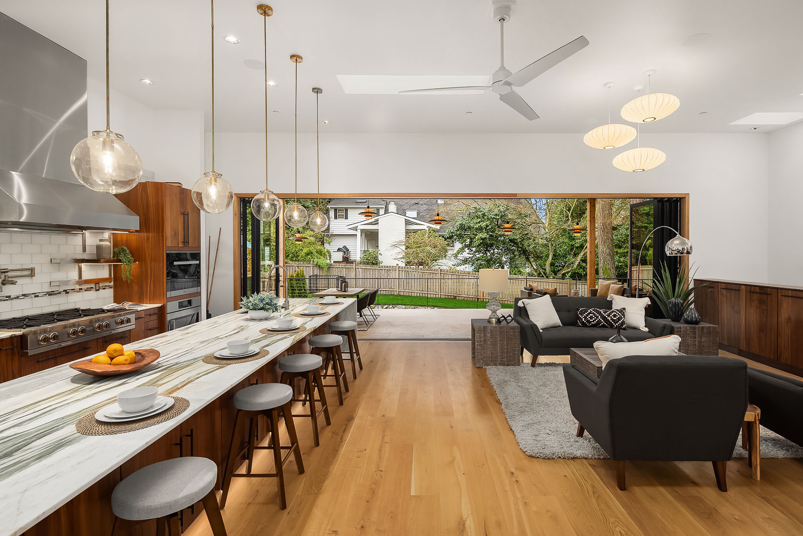 Luxury Mercer Island Mid-Century Modern-19 Modern open-concept kitchen and living area with a marble island, wooden stools, pendant lights, and sliding doors opening to a patio and yard with trees and neighboring houses in the background.