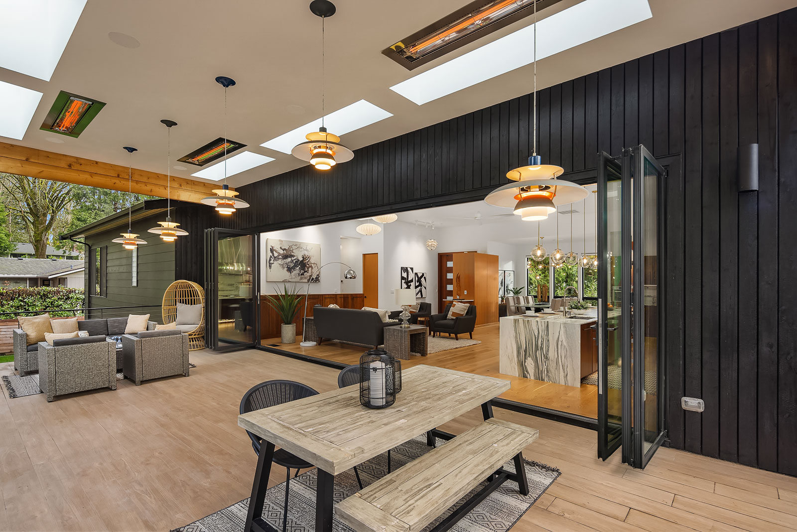 Luxury Mercer Island Mid-Century Modern-20 Modern open-plan living space with folding glass doors connecting a stylish indoor living area and kitchen to an outdoor patio featuring a wooden dining table, lounge chairs, and hanging lights. Skylights provide natural light.