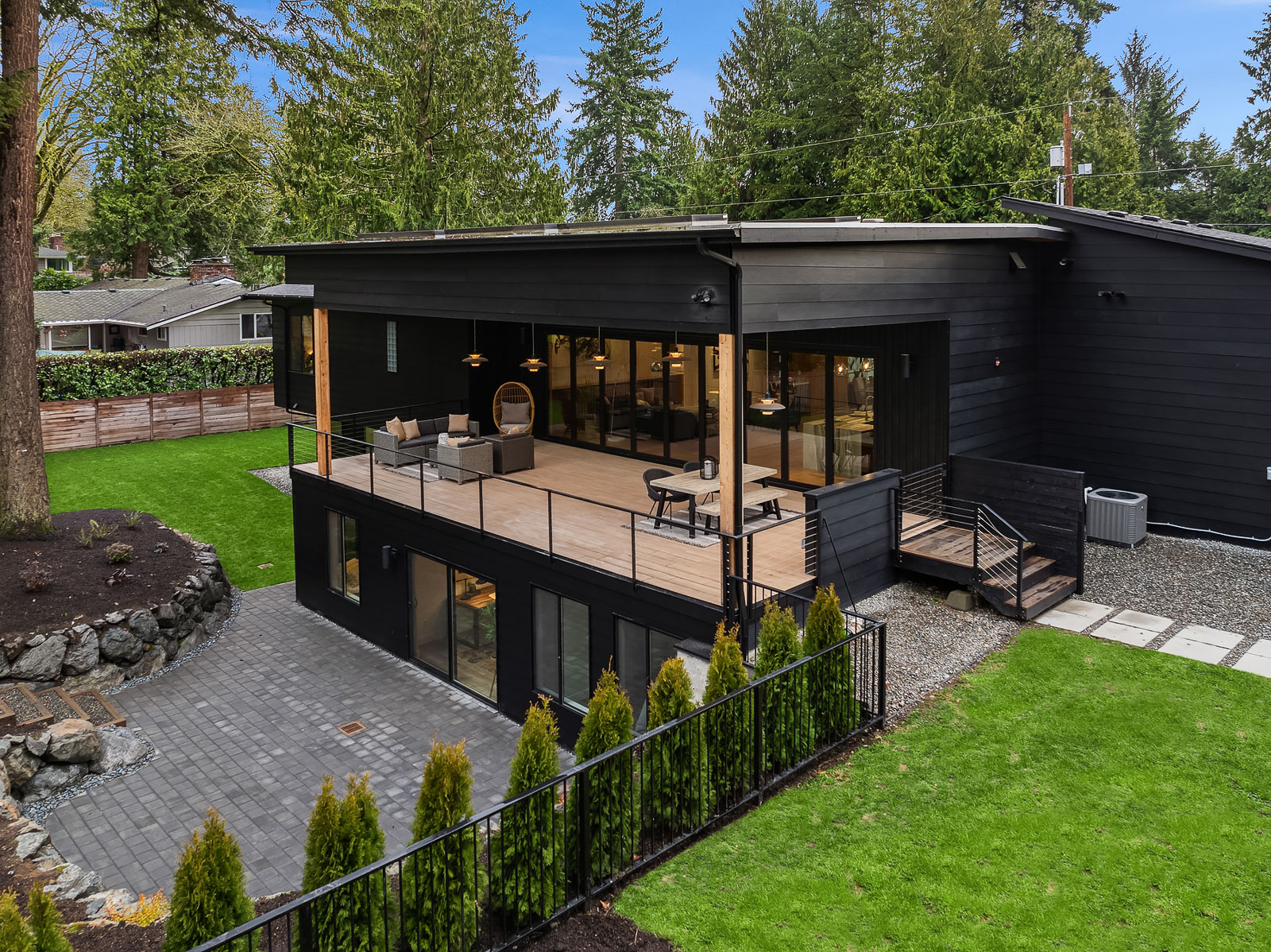 Luxury Mercer Island Mid-Century Modern-22 A modern black two-story house with large windows, a spacious upper deck with outdoor seating, a lower patio, and a green lawn bordered by trees and shrubs.