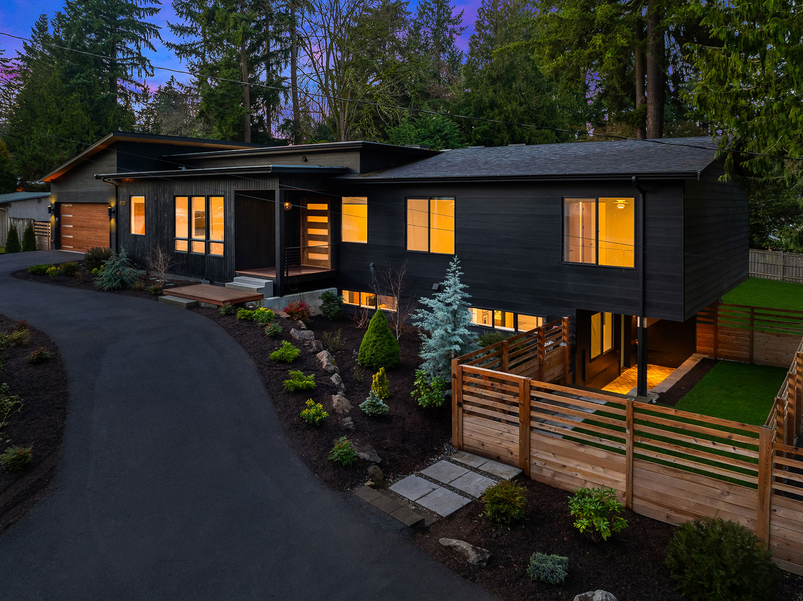 Luxury Mercer Island Mid-Century Modern-5 Modern two-story black house with large windows glowing with warm light at dusk, surrounded by landscaped gardens, a wooden fence, and tall trees; paved driveway curves in front.