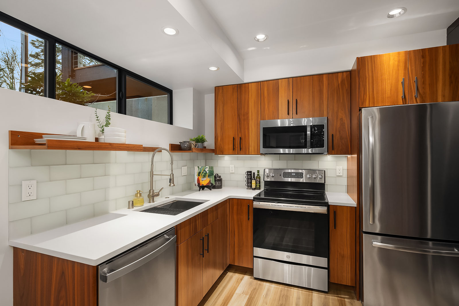 Luxury Mercer Island Mid-Century Modern-6 Modern kitchen with wood cabinets, stainless steel appliances, white countertops, and a white tile backsplash. There are plants and kitchen items on open shelves, under-cabinet lighting, and a large window bringing in natural light.