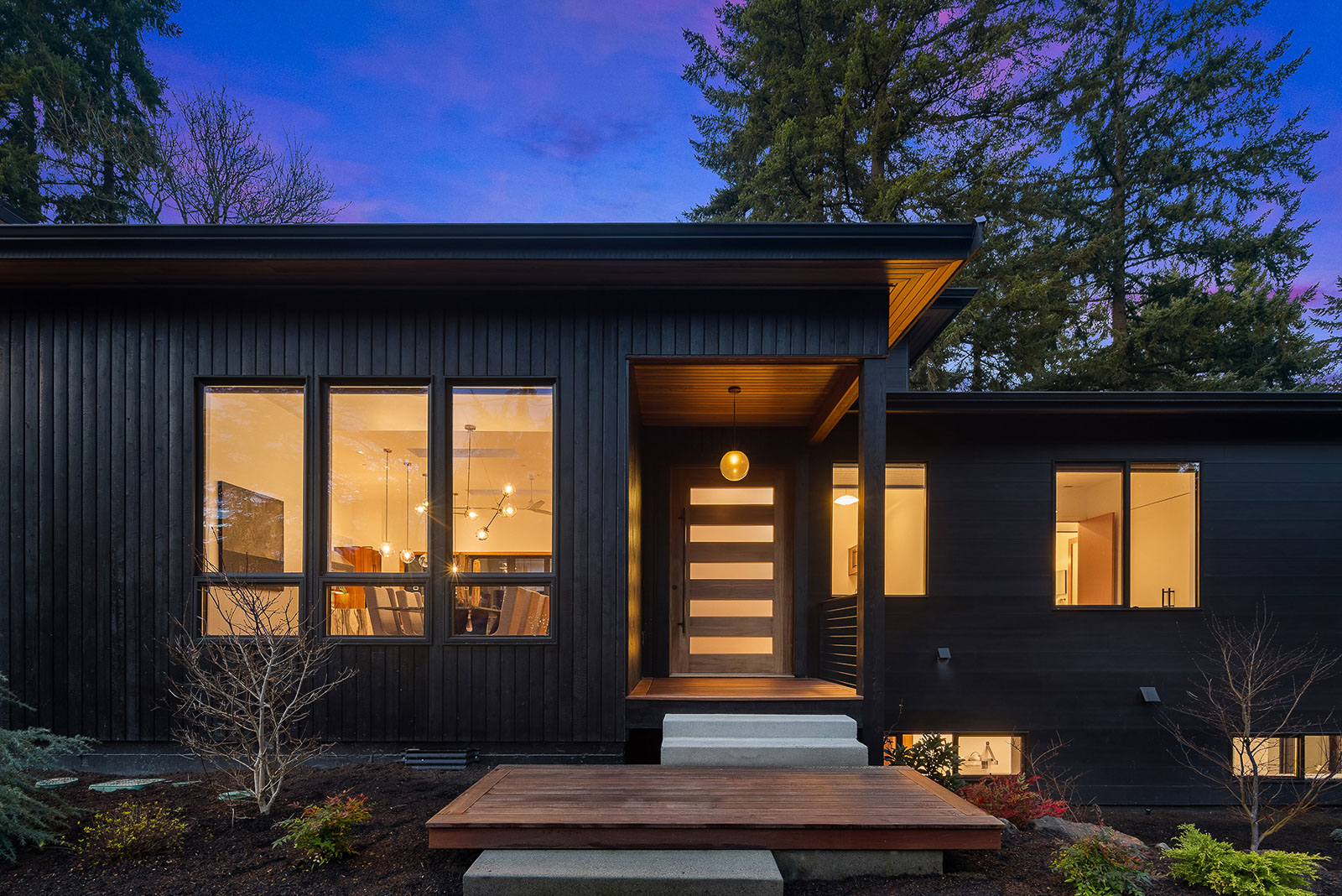 Luxury Mercer Island Mid-Century Modern Modern black house at dusk with large windows lit from inside, a horizontal glass-paneled front door, small porch, and steps leading up, surrounded by trees and landscaped garden.