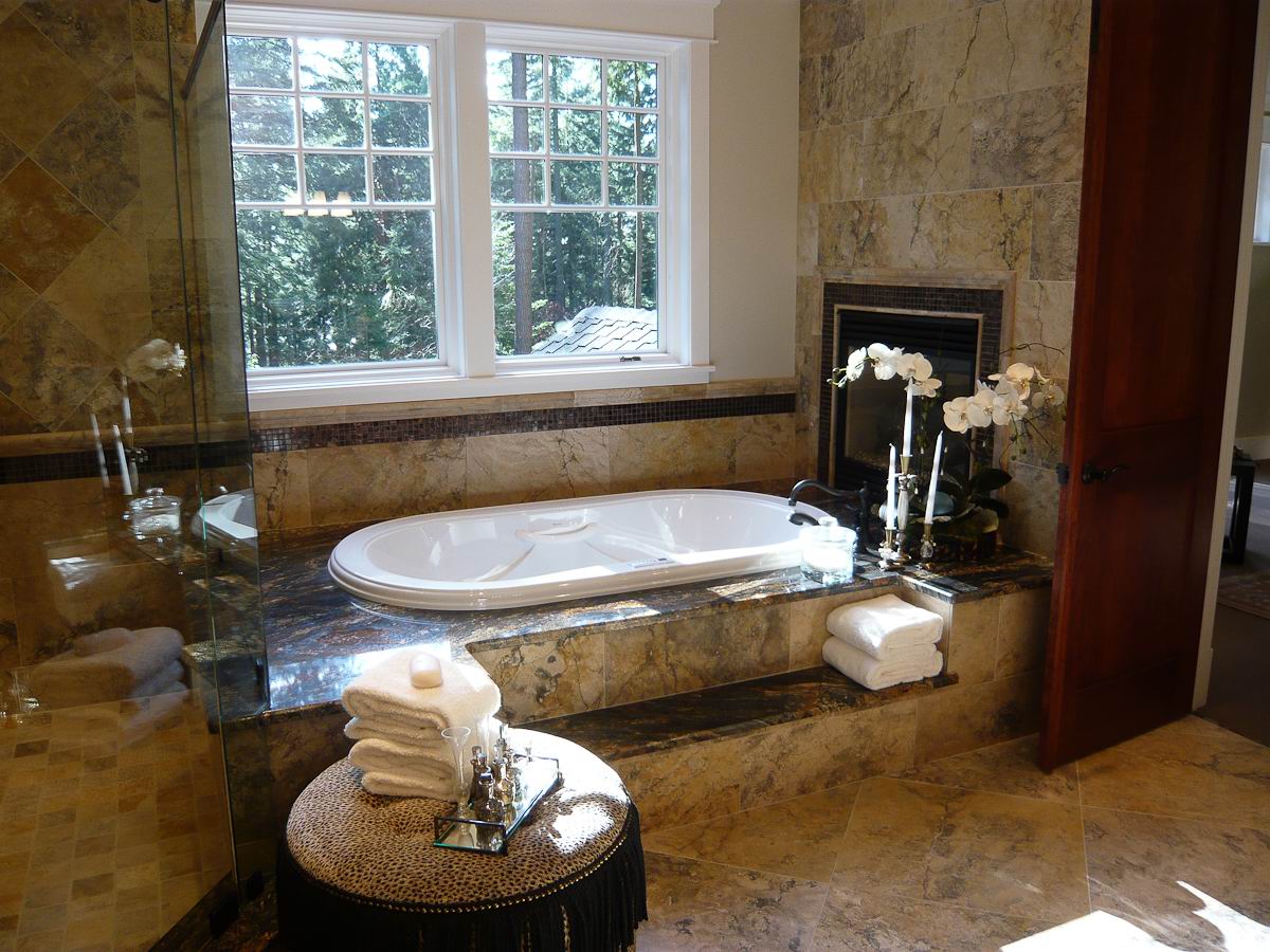 Luxurious bathroom with a built-in bathtub under large windows, marble walls and floor, a glass shower, stacked towels, orchids, and a tray with toiletries on a round ottoman.