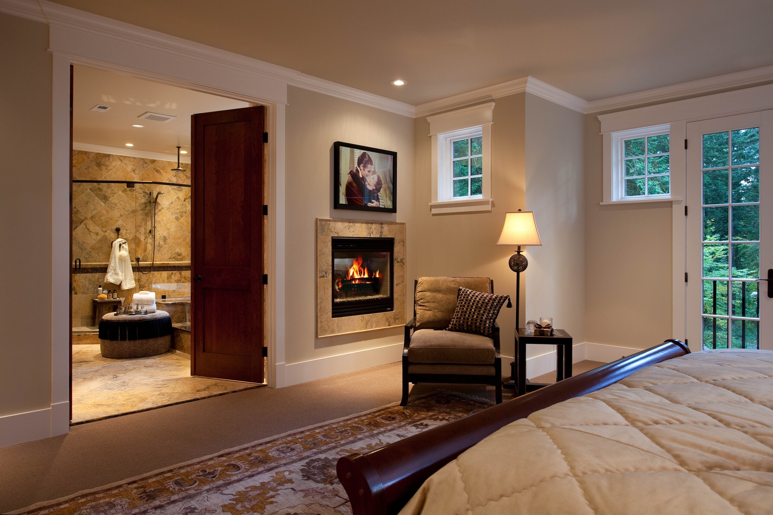 A cozy bedroom with a bed, armchair, lamp, and fireplace. The room connects to a luxurious bathroom with a soaking tub. Large windows let in natural light and offer a view of trees outside.