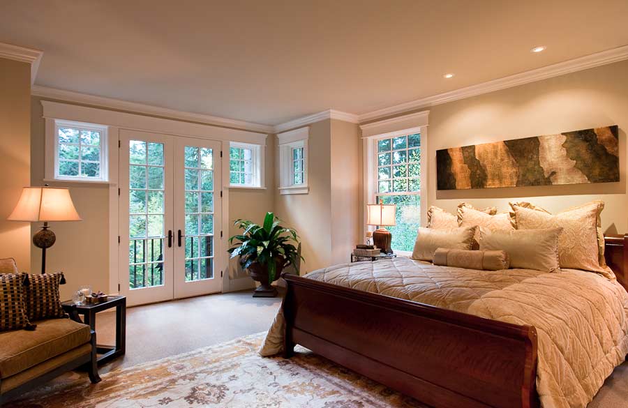 A cozy bedroom with a large wooden bed, beige bedding, an armchair, a floor lamp, potted plant, French doors, windows with white trim, and abstract art above the bed. Warm lighting creates a relaxing atmosphere.