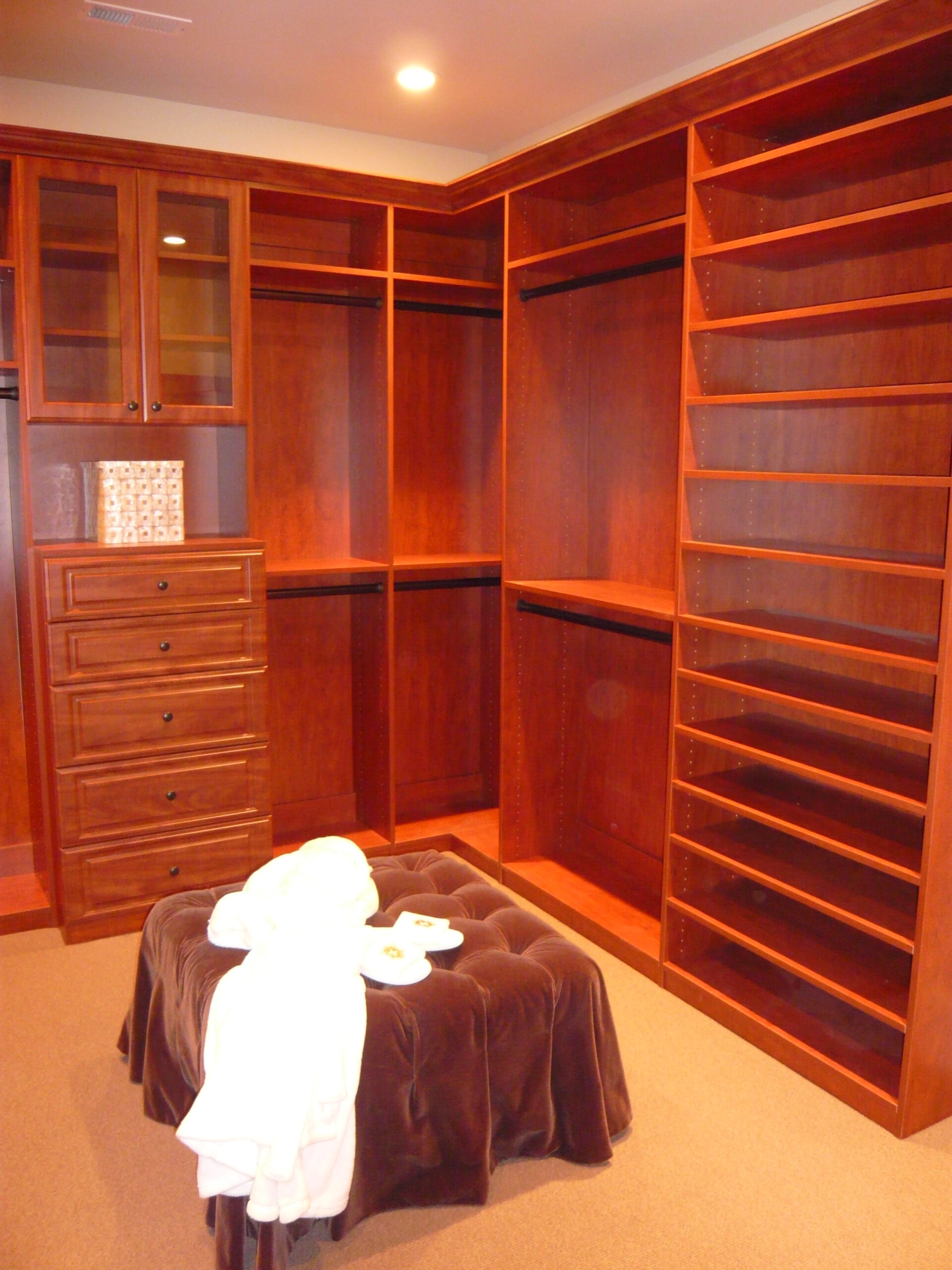 A spacious walk-in closet with wooden shelves, drawers, and hanging rods. A brown ottoman with a white robe and a plate with snacks is in the center. Warm lighting creates a cozy atmosphere.