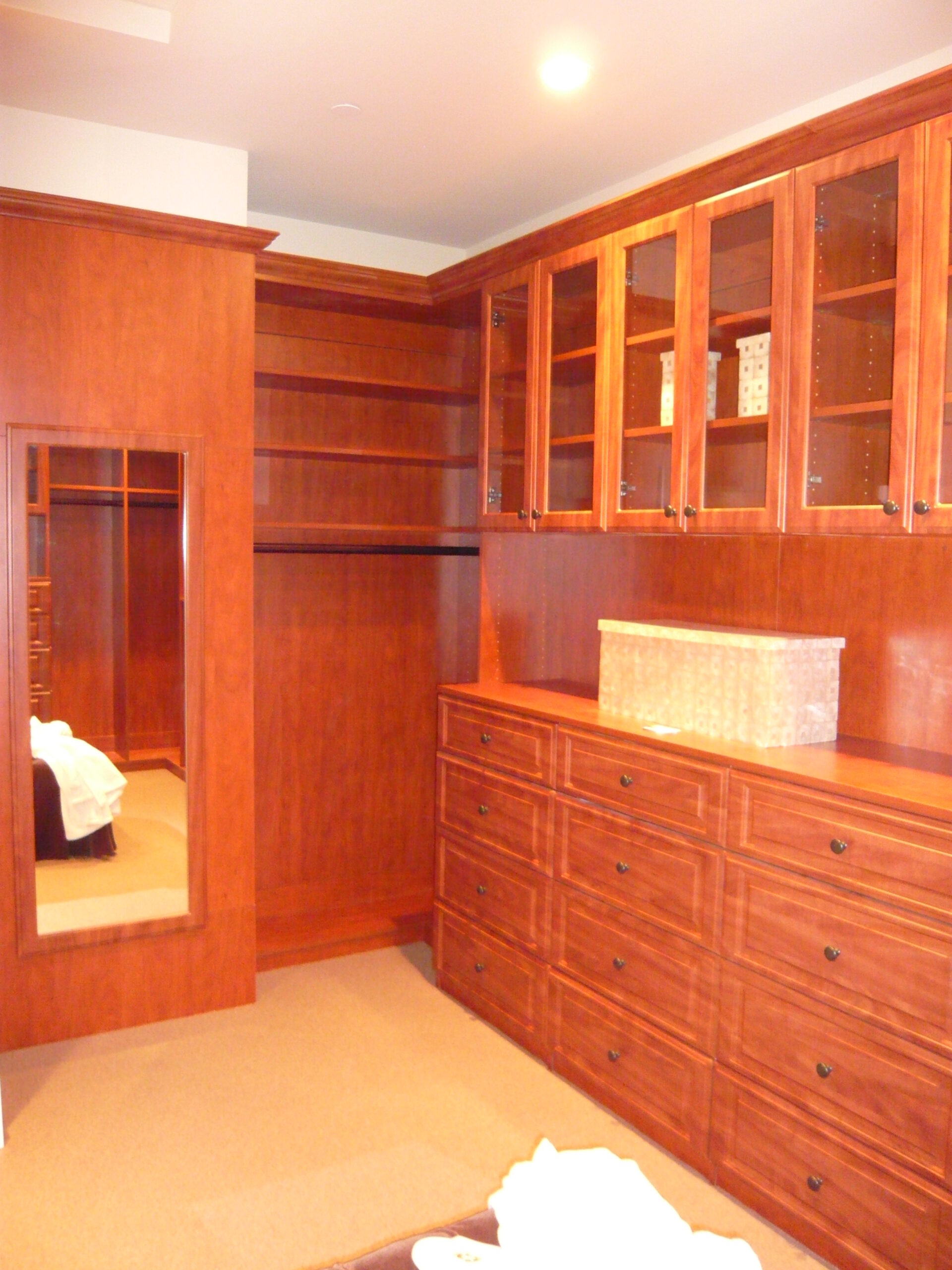 A spacious walk-in closet with wooden shelves, glass-front cabinets, and multiple drawers. A large mirror is mounted on the left wall, and a storage basket sits on the dresser. The floor is carpeted.