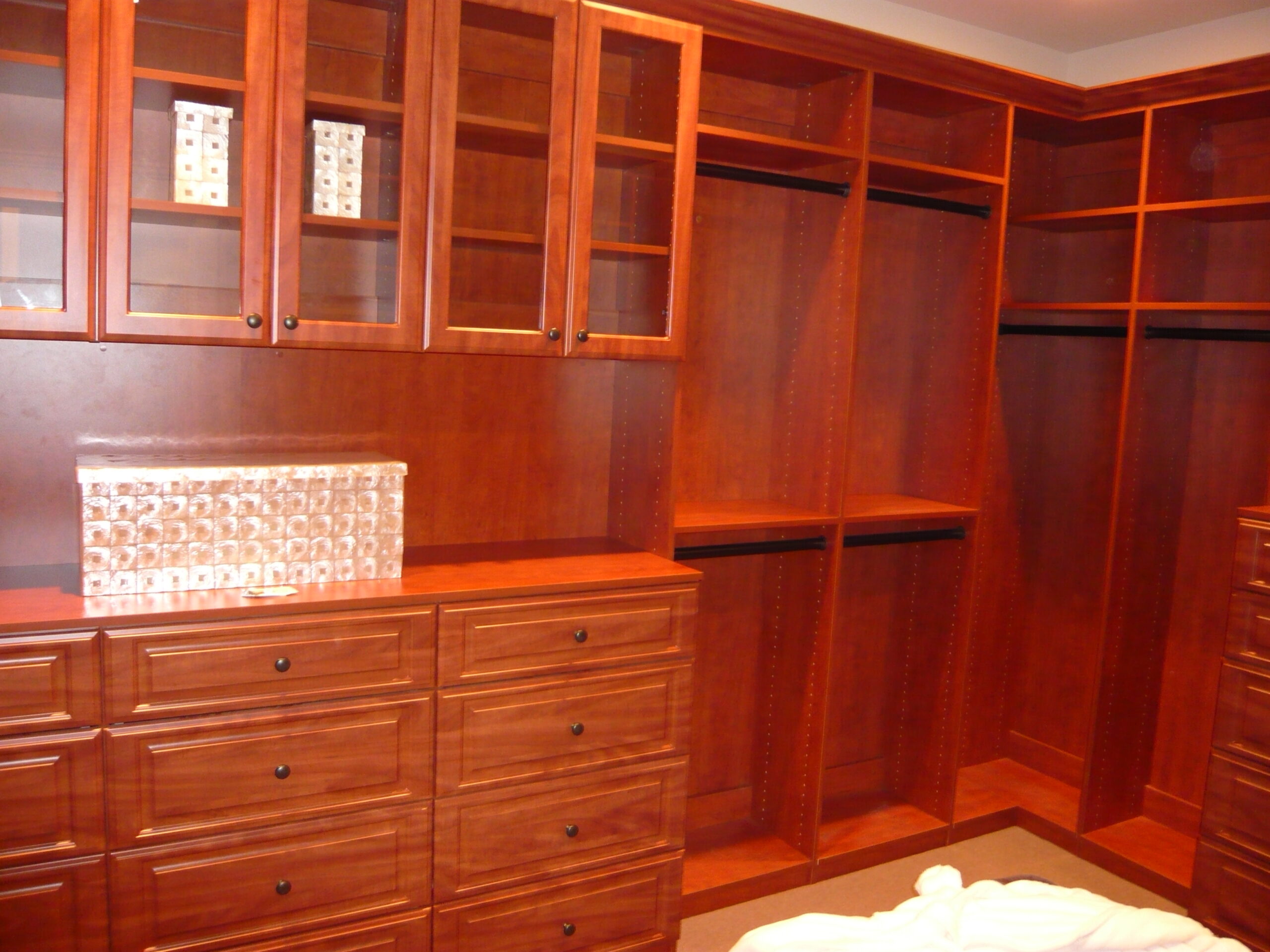 A spacious walk-in closet with wooden cabinetry, drawers, glass-front cabinets, open shelves, and hanging space. Two decorative boxes are displayed on the shelves.