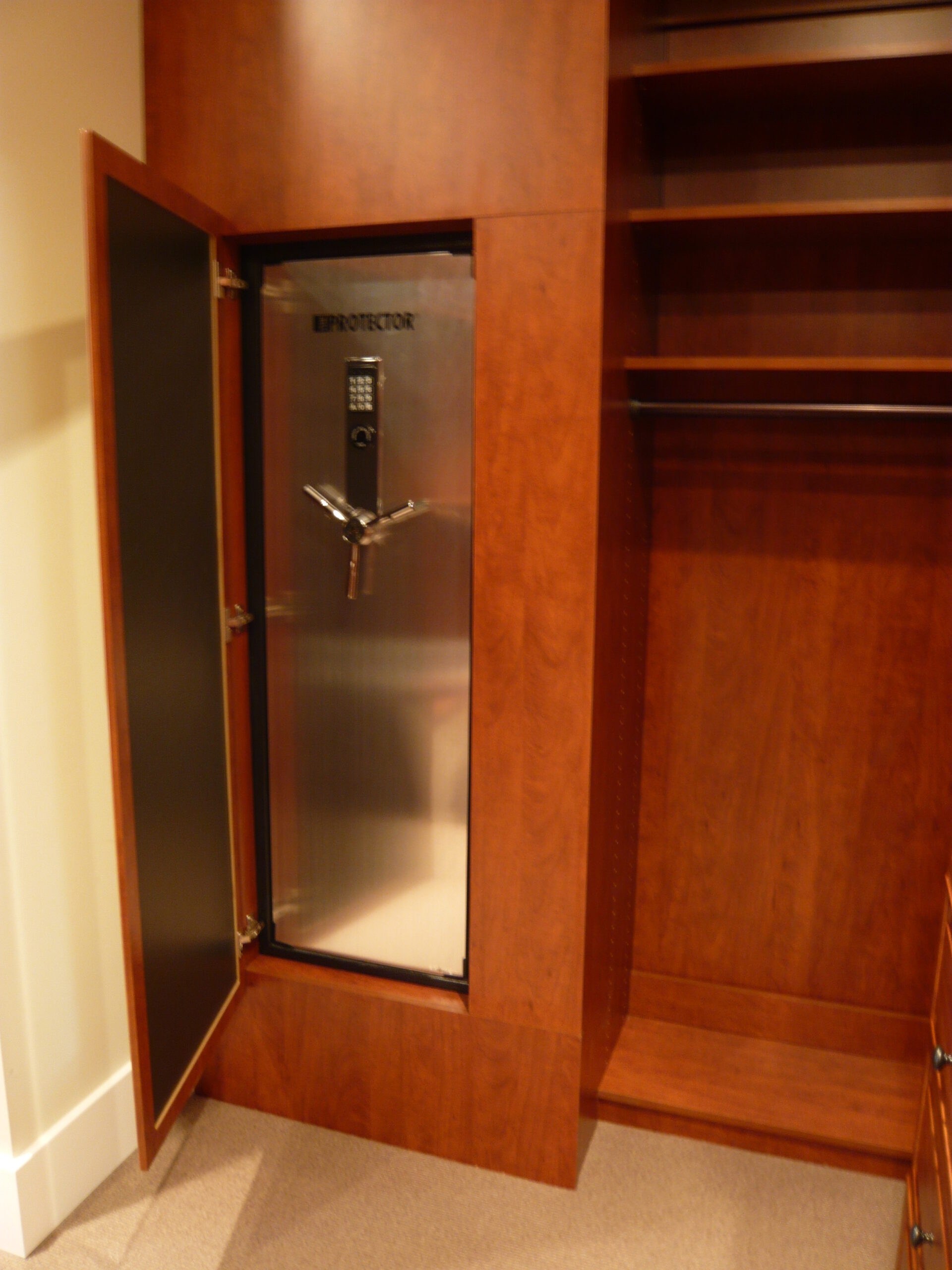 A large safe with a digital keypad and handle is built into a wooden closet. The safe door is open, revealing an empty interior. The closet has shelves and hanging space beside the safe.