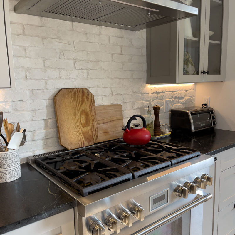 A modern kitchen stove with a stainless steel oven, a red kettle on the burner, wooden cutting boards, utensils in a holder, a toaster, and a white brick backsplash under a metal range hood.