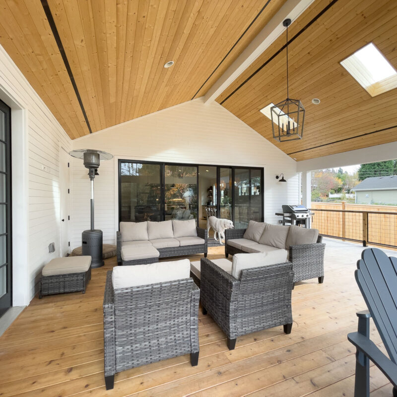 A covered patio with wood ceiling, wicker furniture with beige cushions, a patio heater, a grill, and a dog standing near glass doors; an outdoor area with a fence and greenery is visible.