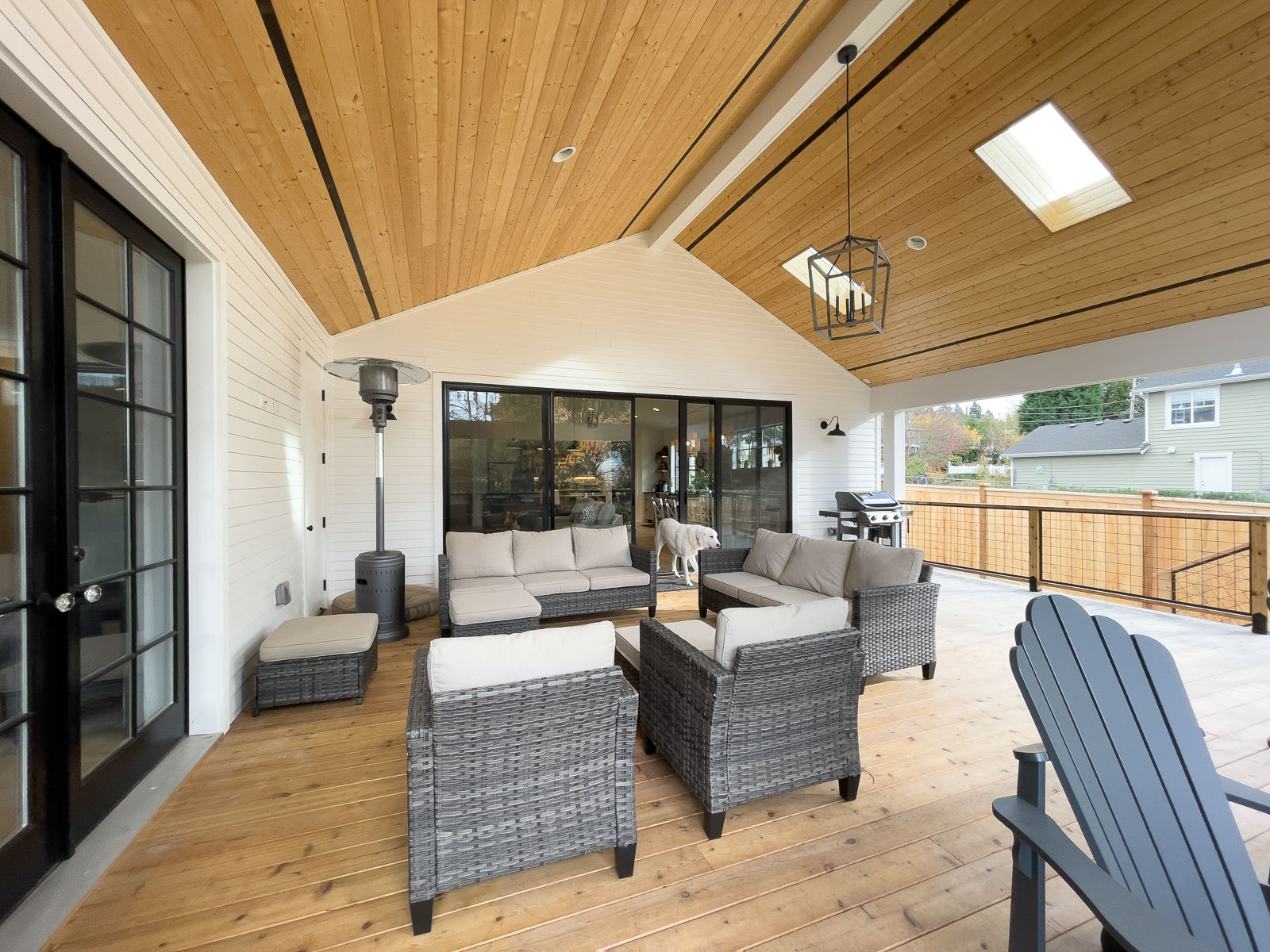 Phillips project Kirkland A covered patio with wood ceiling, wicker furniture with beige cushions, a patio heater, a grill, and a dog standing near glass doors; an outdoor area with a fence and greenery is visible.
