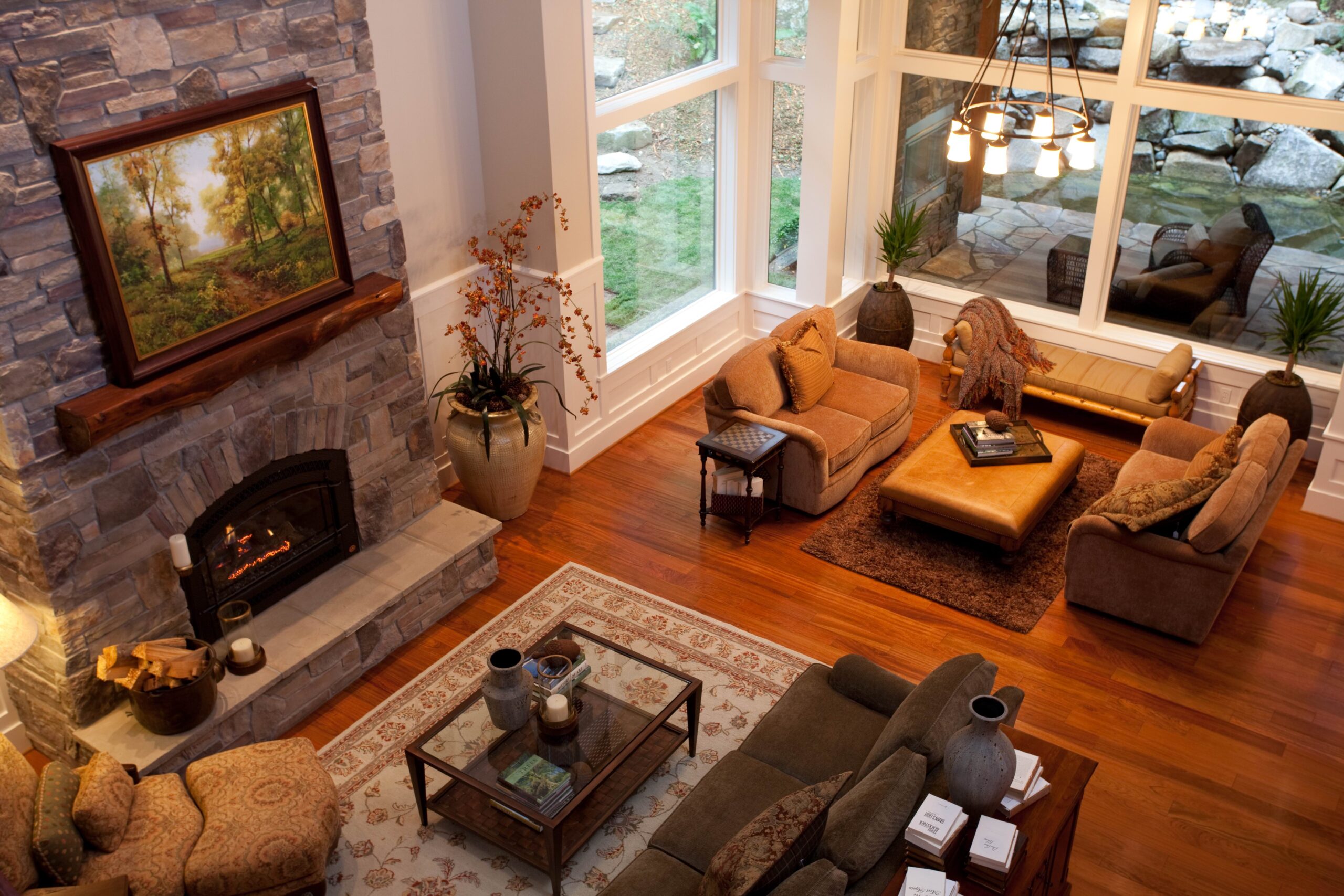A cozy living room with wooden floors, a stone fireplace, large picture windows, comfortable seating, a coffee table, and decor including vases, books, and a landscape painting above the fireplace.
