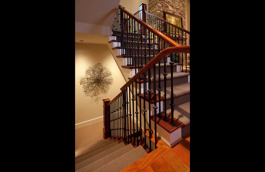 A view of a carpeted staircase with wooden handrails and black iron balusters, featuring a stone accent wall and artwork at the top landing and a decorative metal wall sculpture on the side wall.