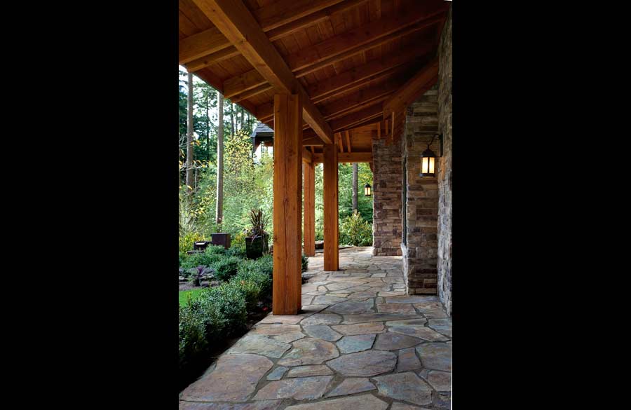 A rustic stone walkway leads under a wooden porch roof supported by thick timber posts, with stone walls and outdoor lanterns, surrounded by lush greenery and trees.