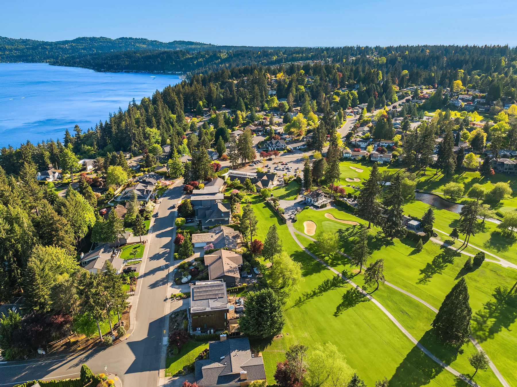 Aerial view of a suburban neighborhood with green lawns, trees, houses, and a golf course near a large blue lake, surrounded by forested hills under a clear blue sky.
