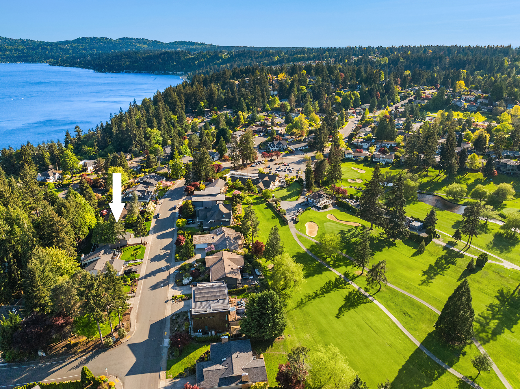 Aerial view of a suburban neighborhood next to a golf course and a lake, surrounded by tall trees. A white arrow points to a specific house in the neighborhood. The weather is clear and sunny.