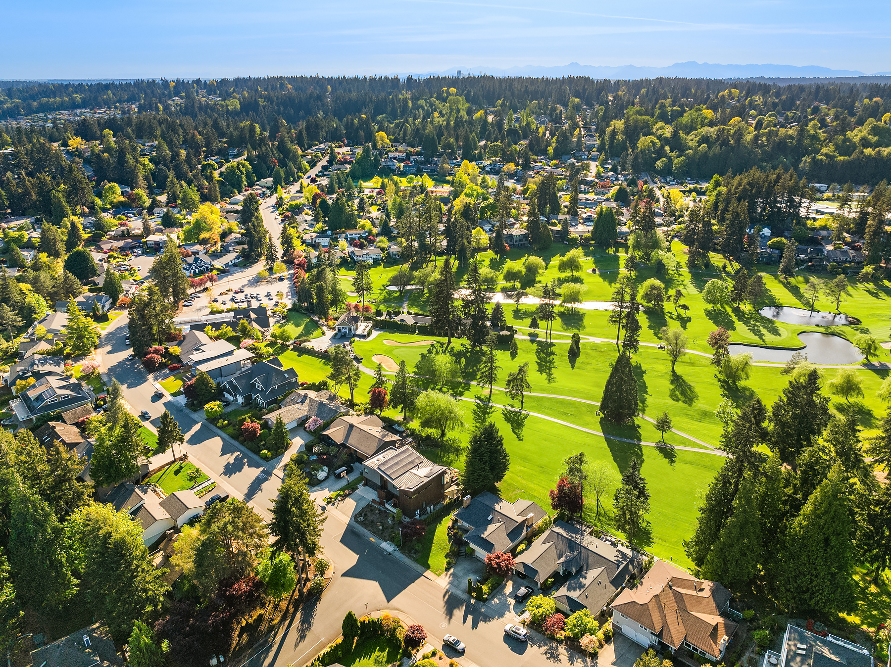 Aerial view of a suburban neighborhood with houses surrounded by trees, next to a large, green golf course with sand bunkers, under a clear blue sky.