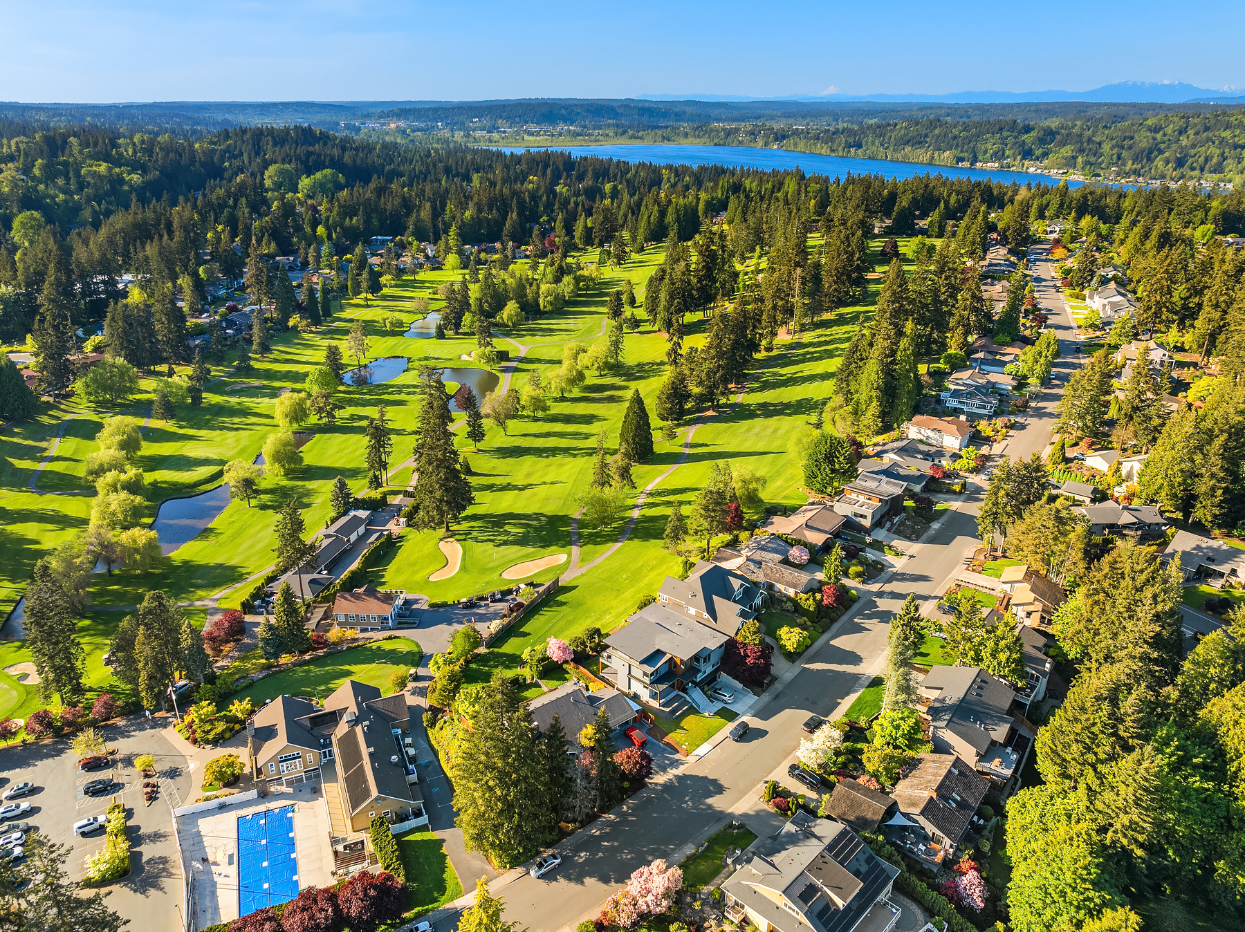 Aerial view of a suburban neighborhood with houses lining a street next to a large, green golf course with sand traps, ponds, and trees, set near a lake and surrounded by forests under a clear blue sky.